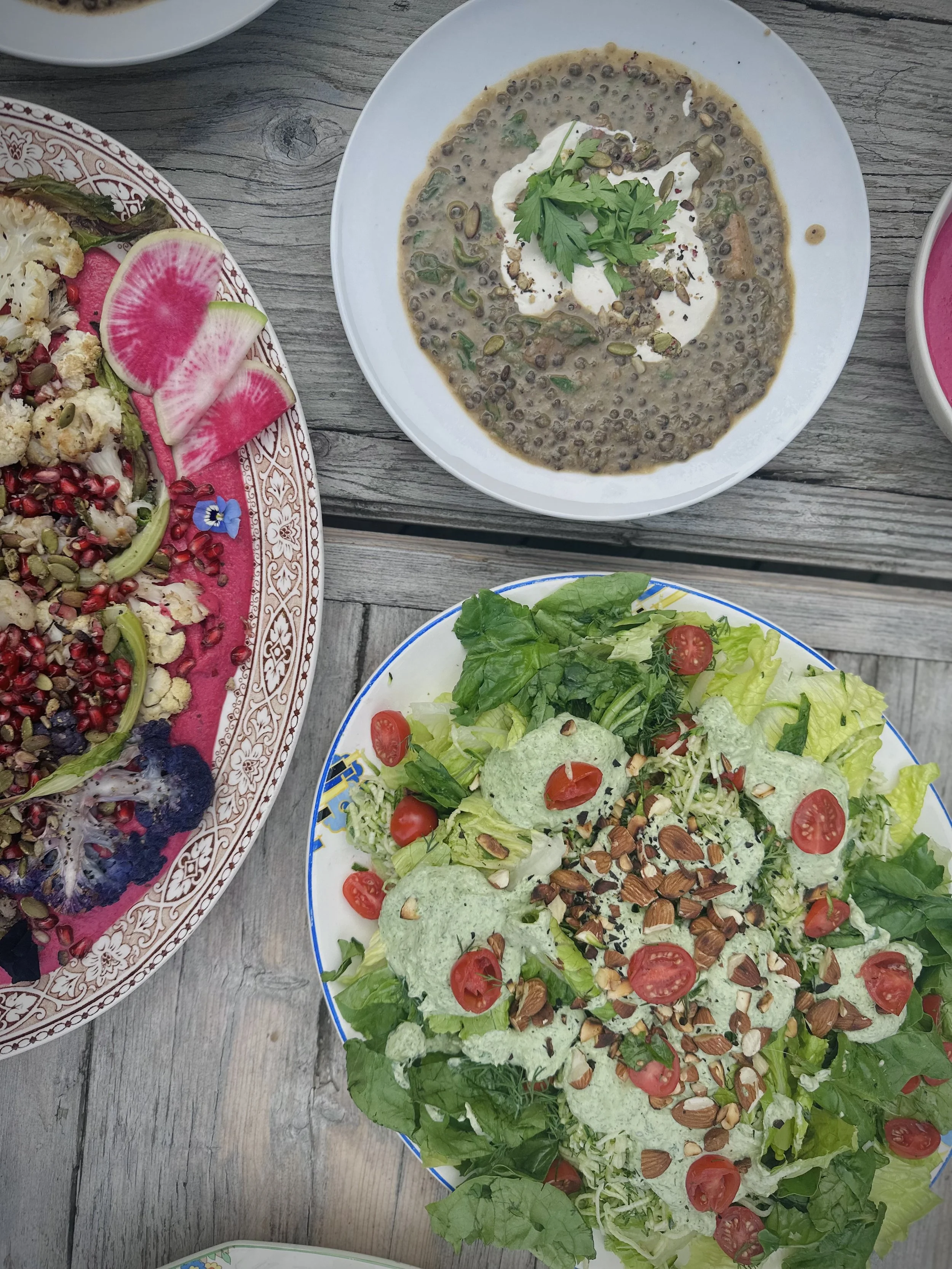 Assorted Mediterranean dishes including a lentil soup garnished with herbs and yogurt, a fresh green salad with cherry tomatoes, chopped almonds, and green dressing, and a roasted vegetable platter with radish slices.