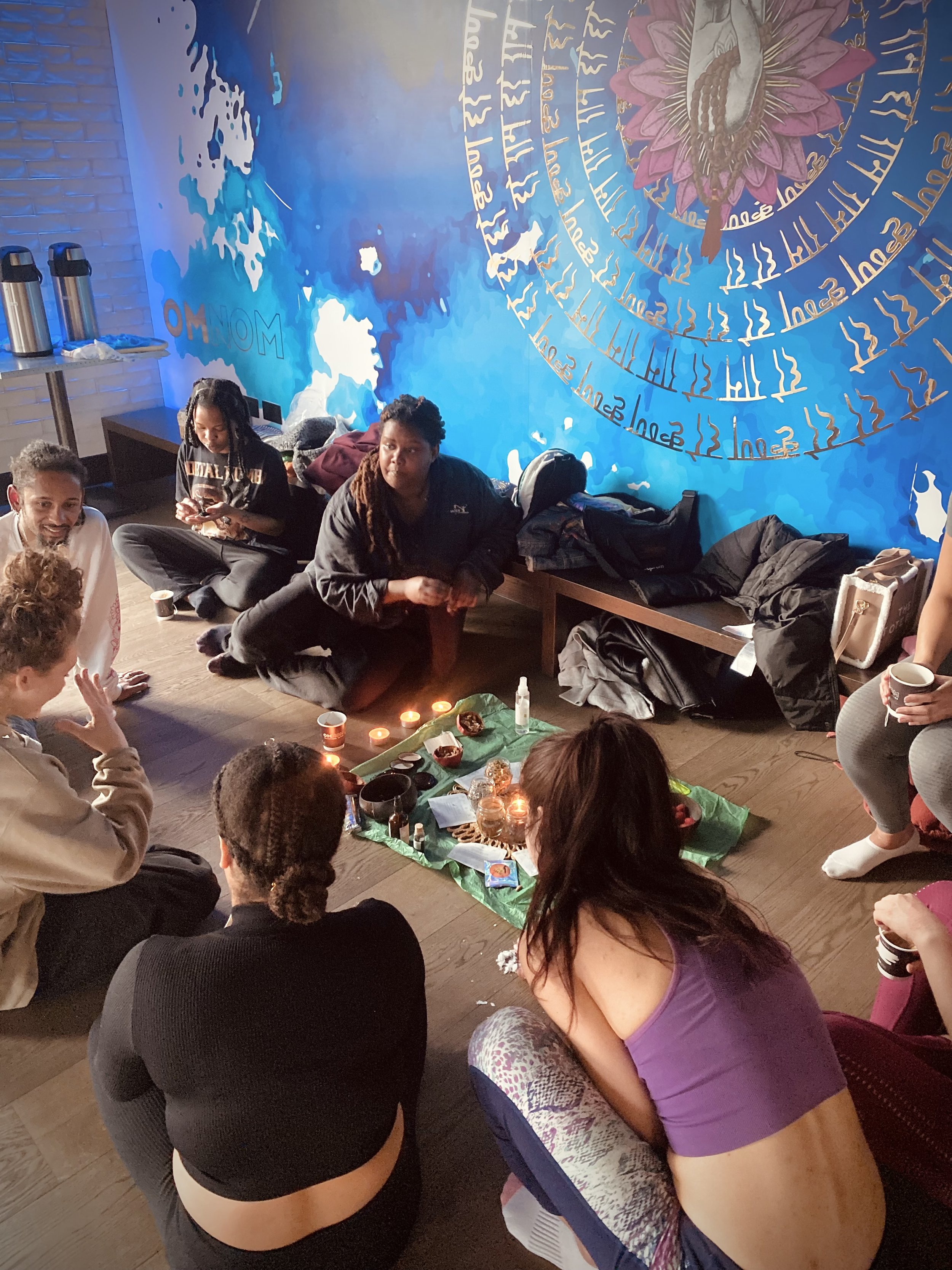 A group of people sitting in a circle on the floor in a room with a blue and white painted wall and a large mural. They are engaged in a spiritual or meditation practice with candles and small bowls in the center.