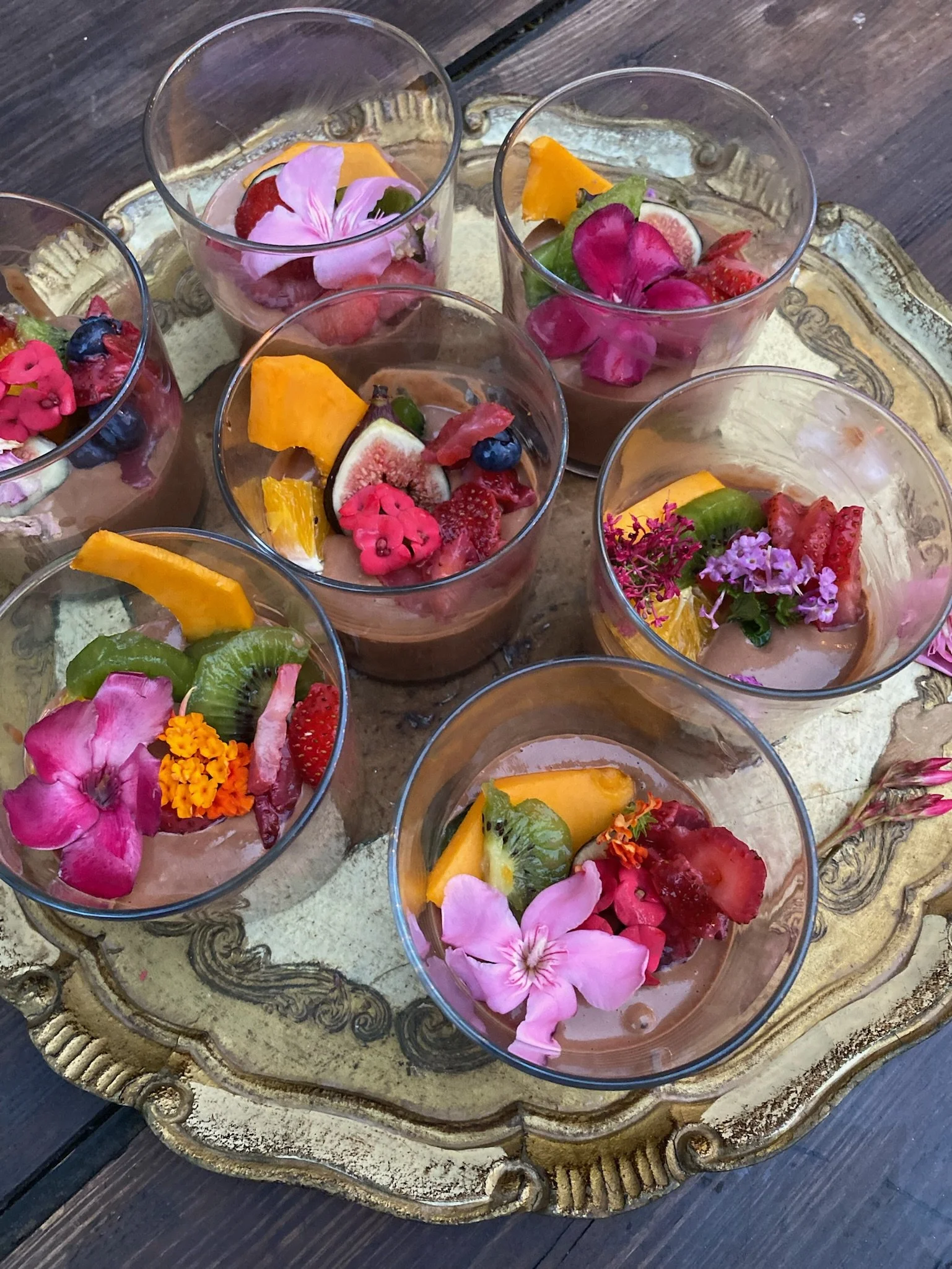 Several glasses of chocolate mousse topped with colorful edible flowers and assorted fresh fruits on an ornate tray.