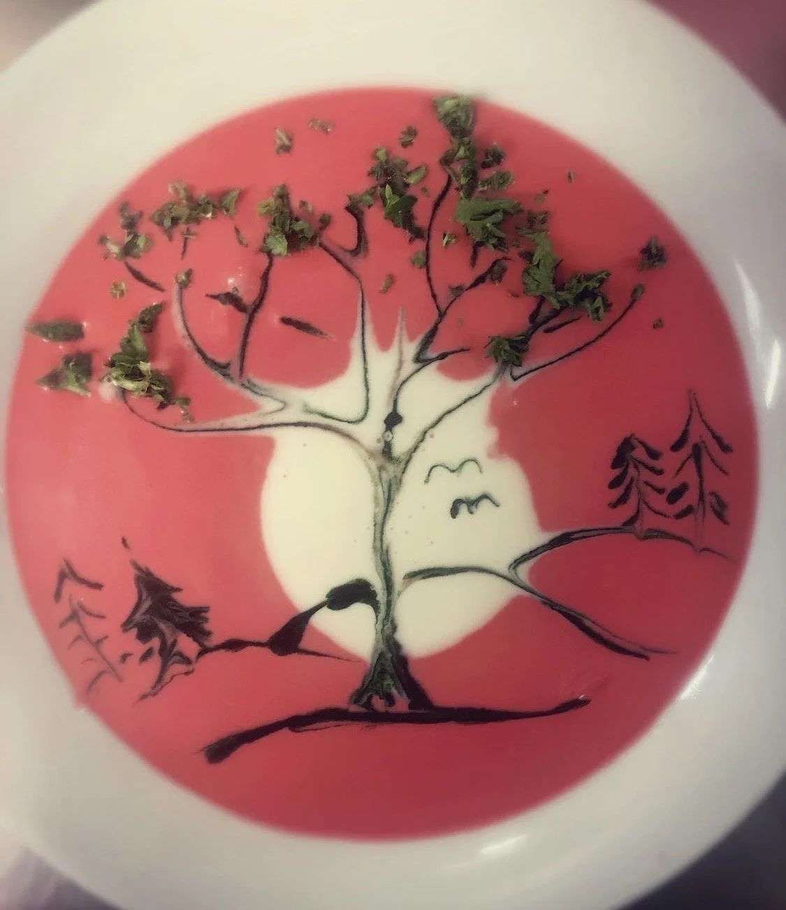 A bowl of pink and white soup with a black line drawing of a tree and mountains, decorated with dried herbs.