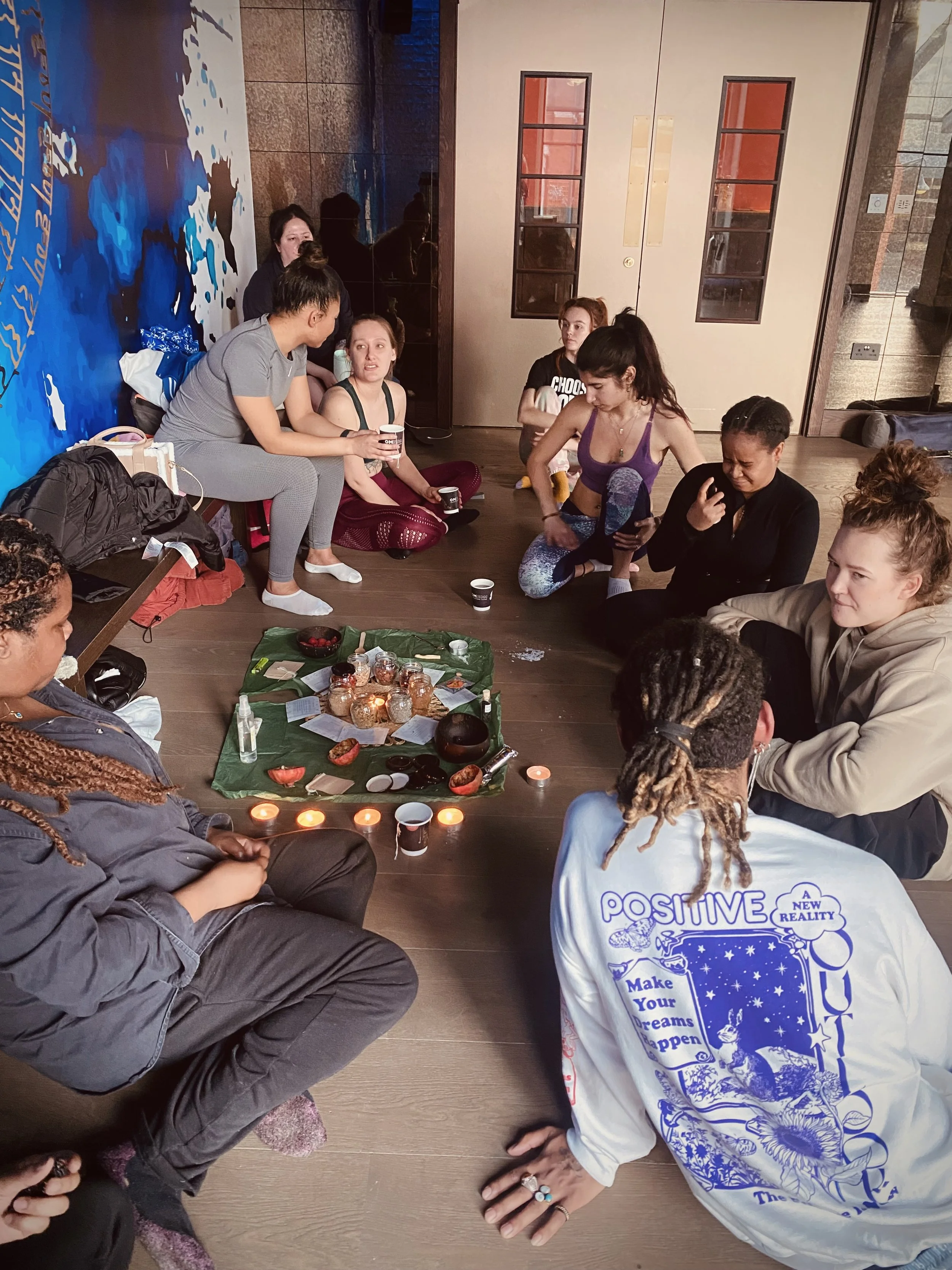 Group of women sitting on the floor in a circle, engaging in a meditation or spiritual practice with a central arrangement of candles, stones, and papers.