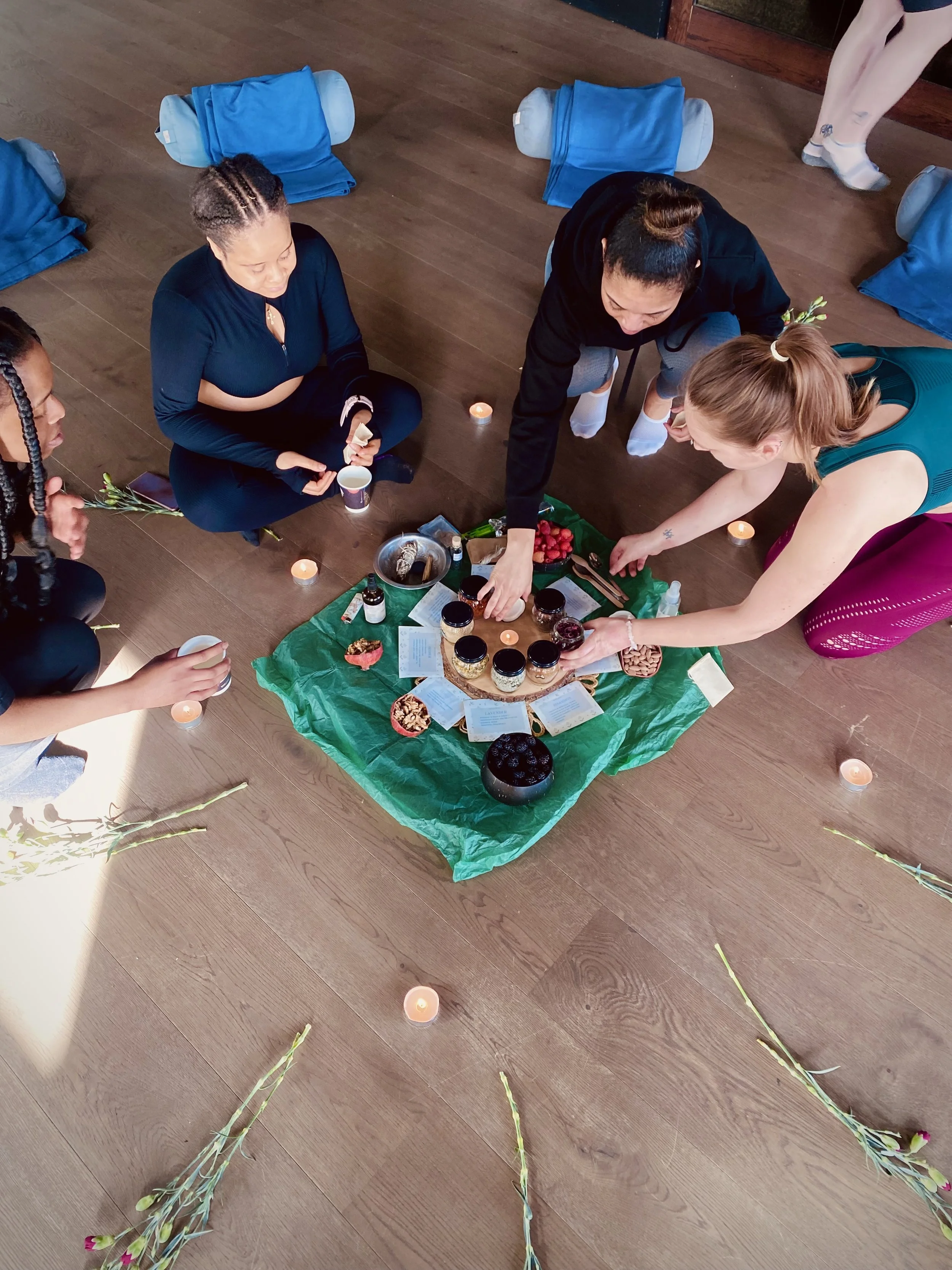 A group of people sitting and kneeling on a wooden floor around a display of jars, bowls, and notes, with small candles and flowers surrounding them.