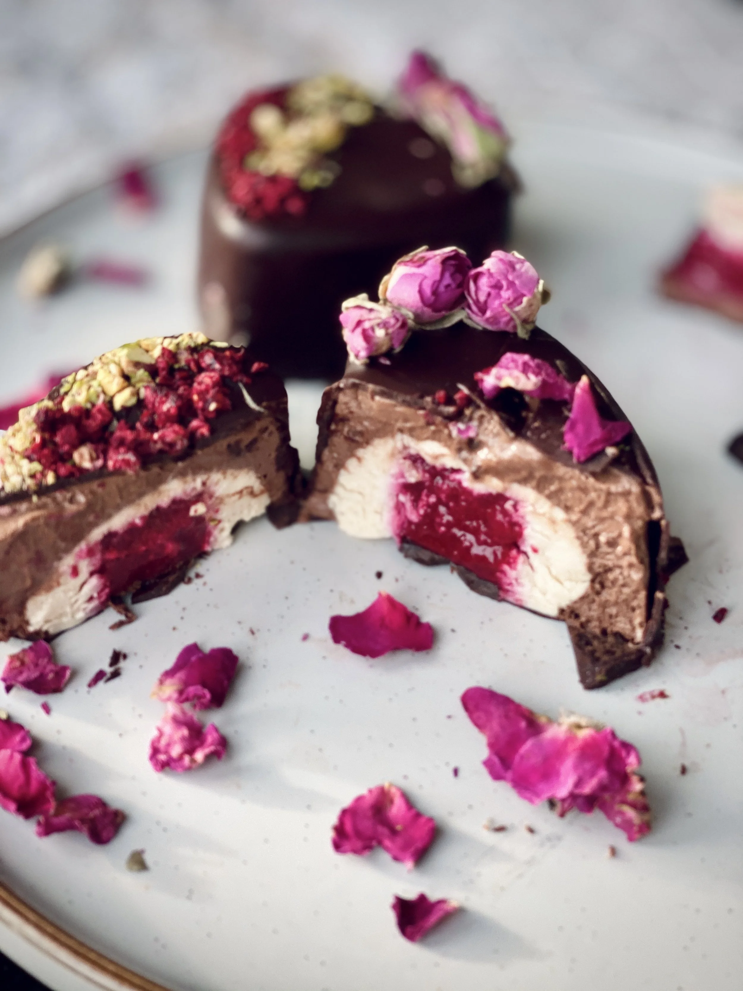 Chocolate dessert with pink rose decorations, cut in half to reveal a cherry filling and creamy center, placed on a white plate with scattered rose petals.