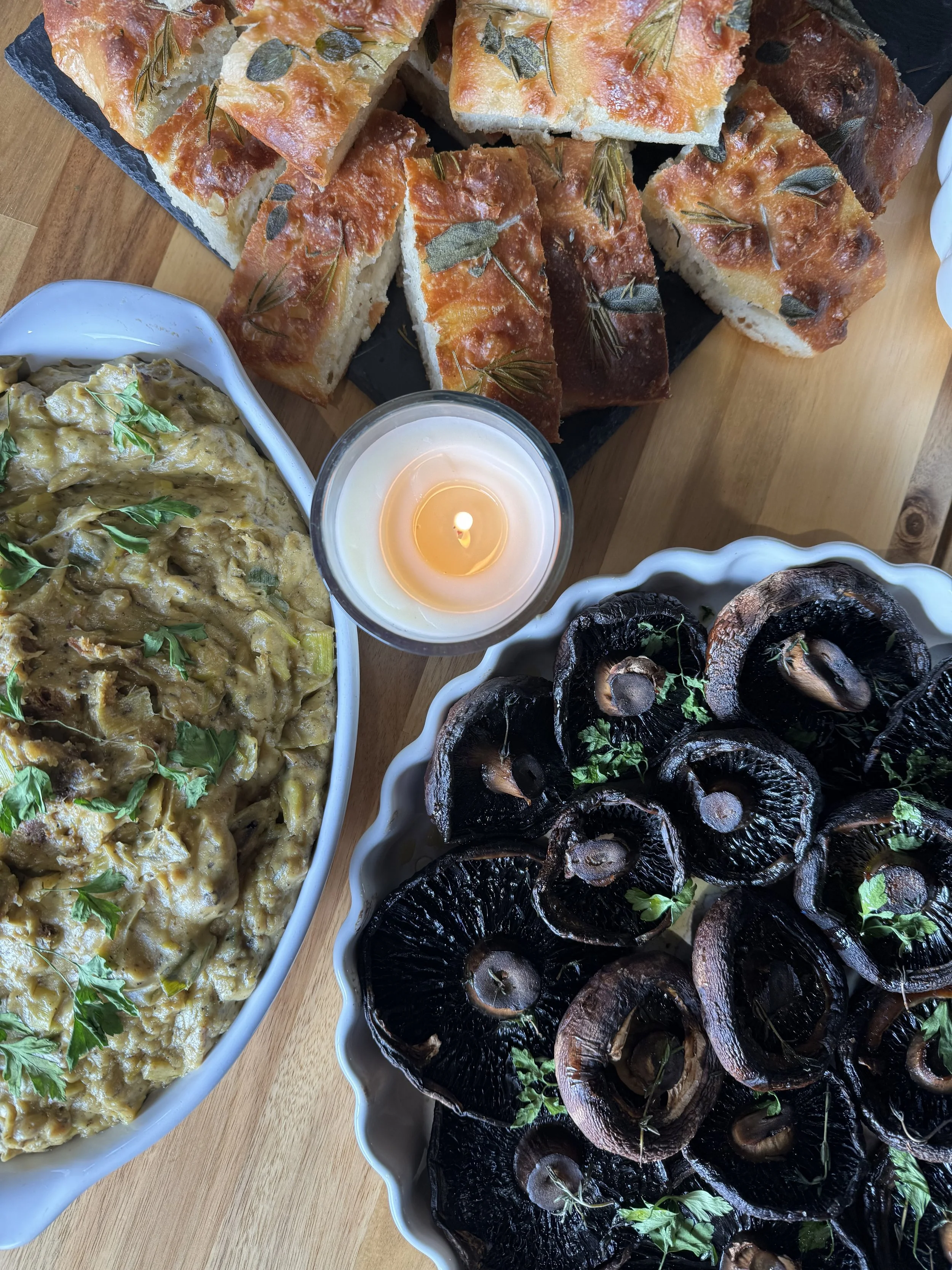 A spread of food on a wooden table, including focaccia bread with herbs, a dish of mushroom ragout, roasted mushrooms topped with herbs, and a lit candle in the center.