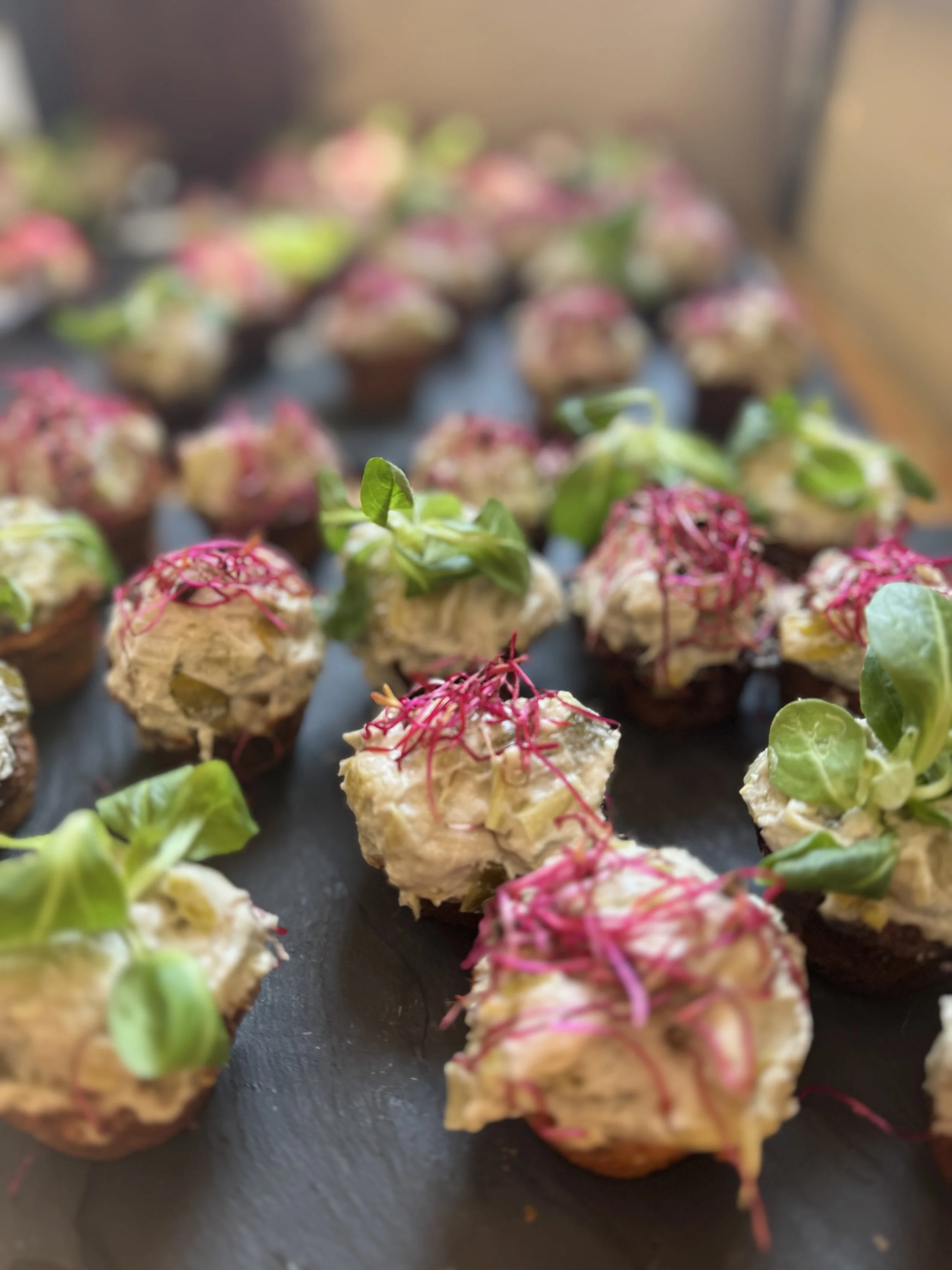 Close-up of bite-sized appetizers with creamy topping, green microgreens, and pink shredded garnish arranged on a black tray.