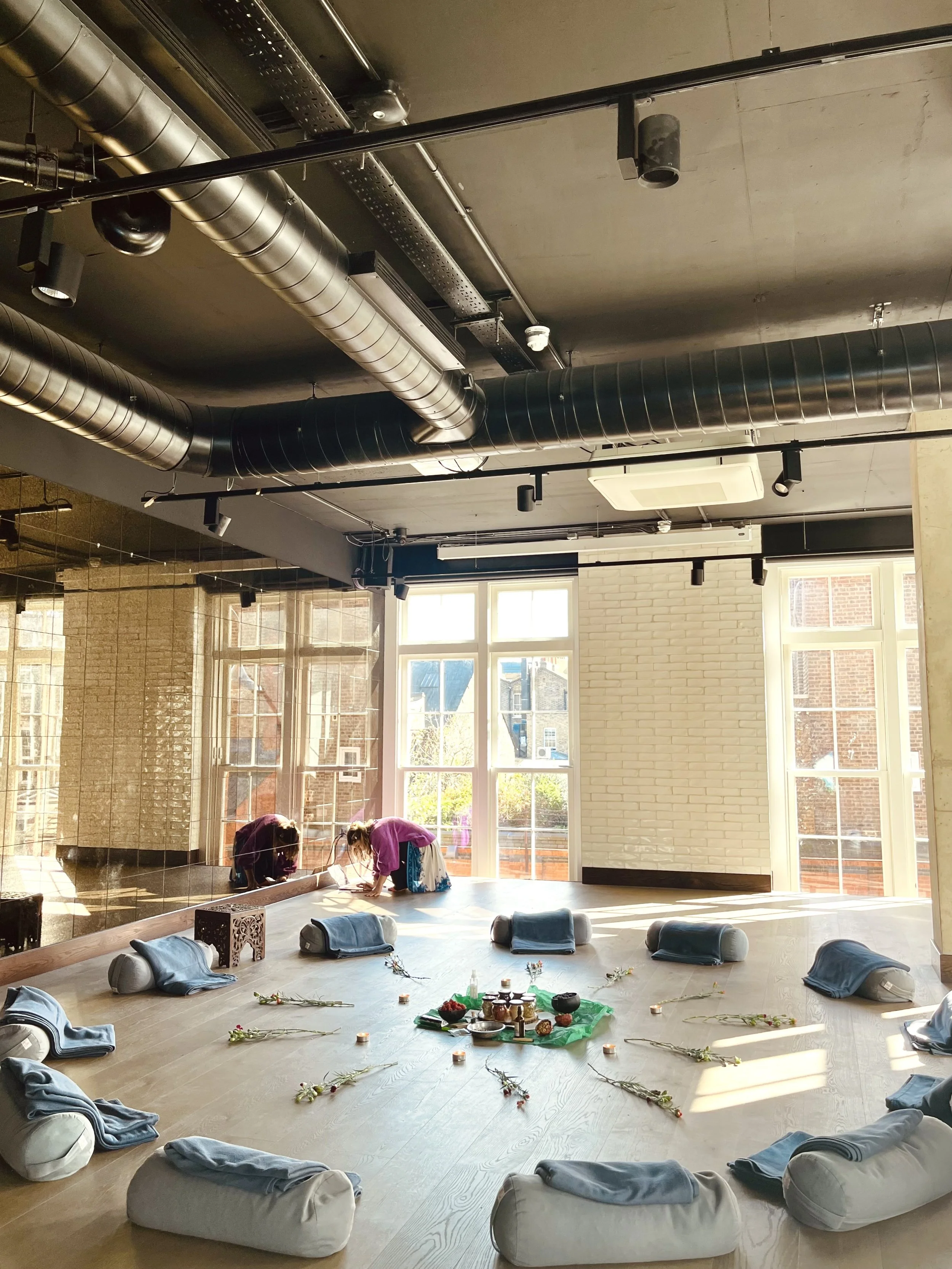 Indoor yoga studio with sunlight streaming through large windows, arranged with floor cushions, flowers, and a green cloth with ritual items, as two women practice yoga or meditation