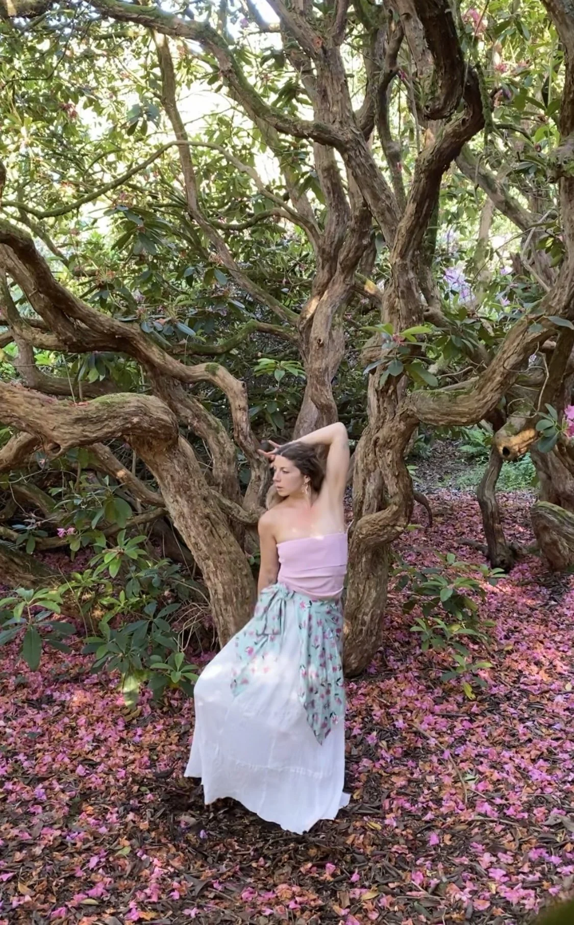 A woman standing among twisted branches of a tree with pink flowers and fallen petals on the ground, wearing a strapless pink top and a flowing white skirt with a floral scarf.