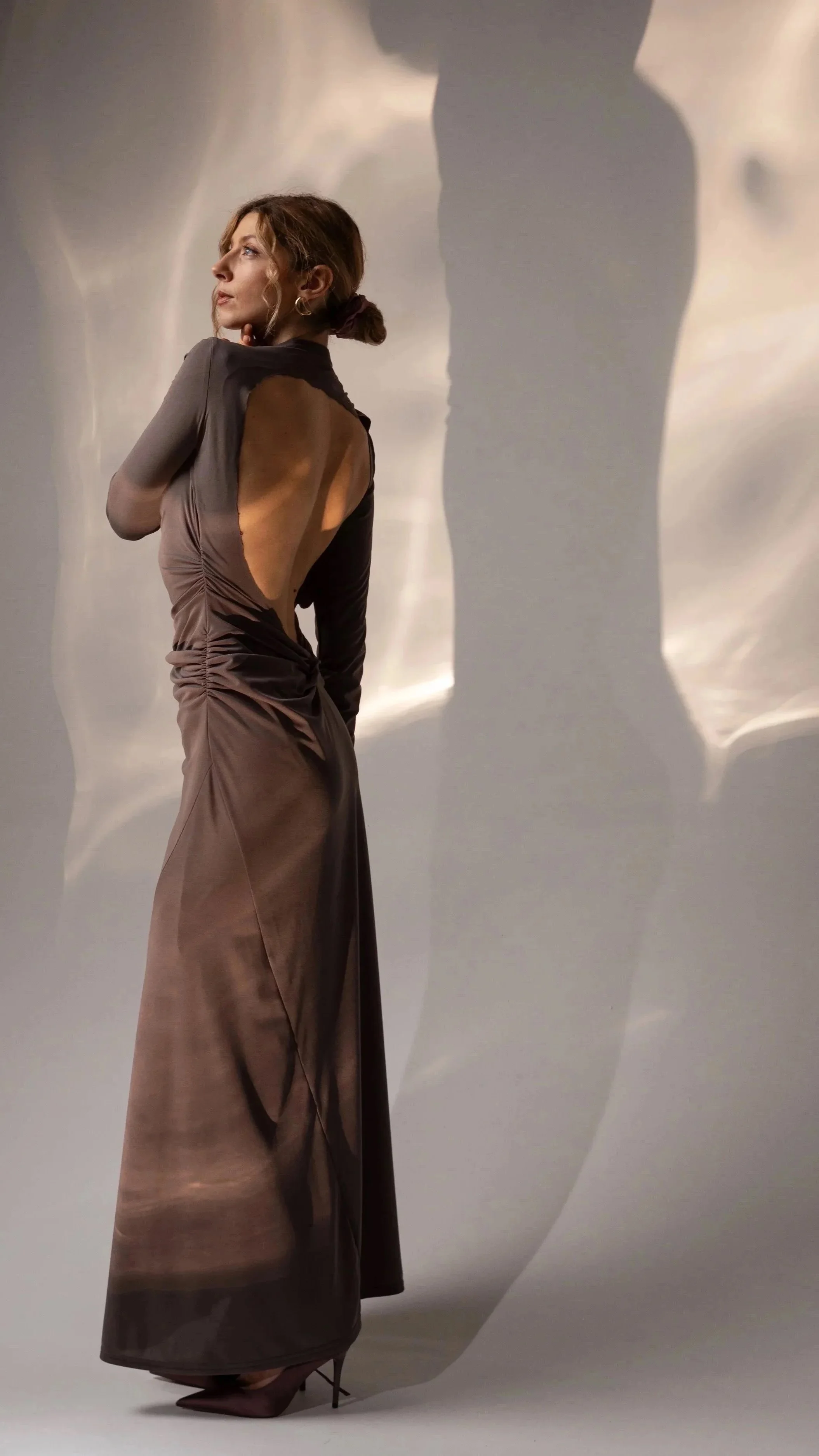 A woman in a long, elegant gown with an open back, standing against a plain background with shadows cast on the wall.