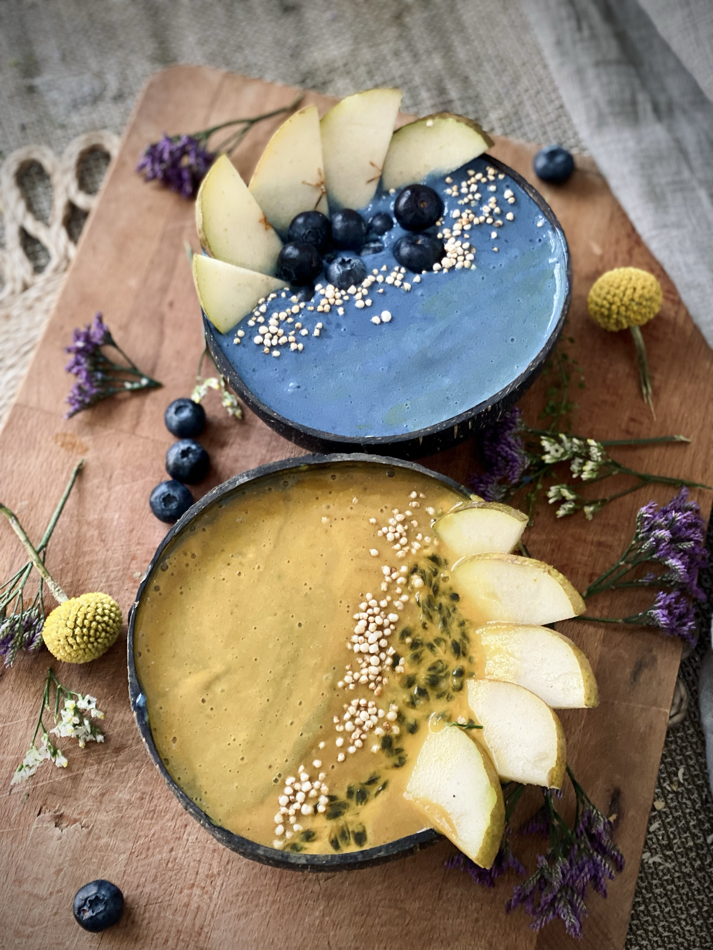 Two vibrant fruit smoothies in halved, decorated with slices of pear, blueberries, passion fruit, and white sprinkles, surrounded by flowers on a wooden tray.