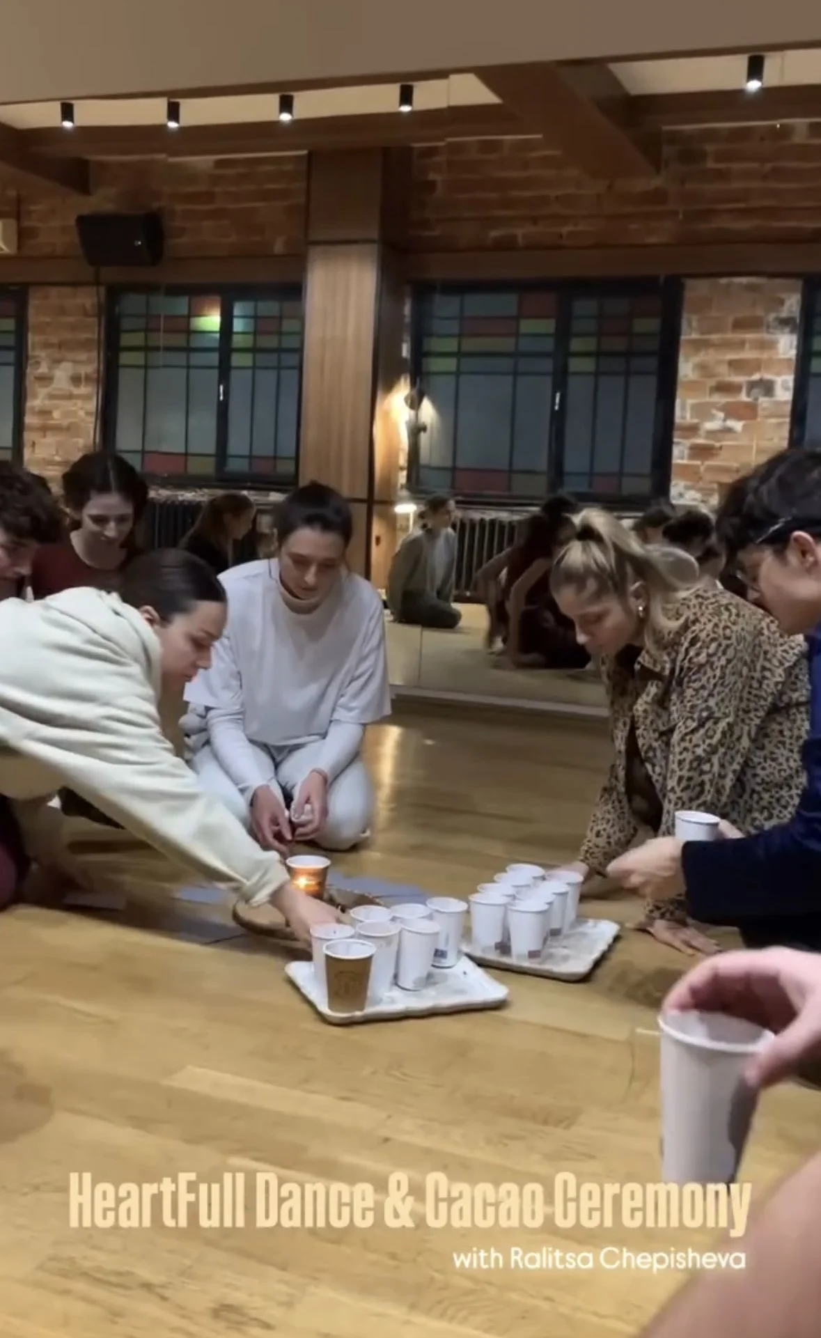 People sitting on the floor participating in a ceremony involving white cups and a candle in a cozy room with brick walls and stained glass windows.