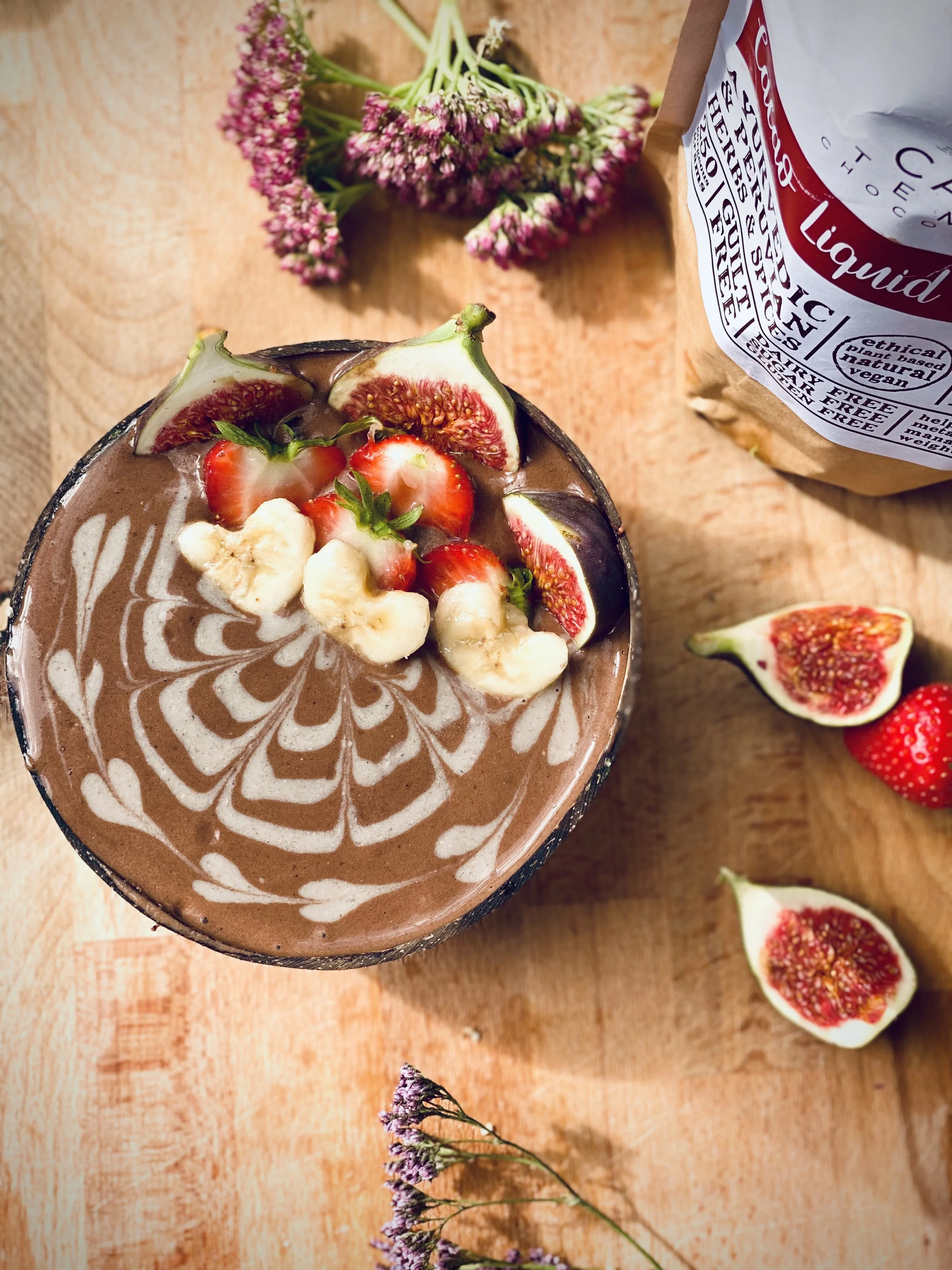 A glass of chocolate drink decorated with white chocolate design topped with sliced strawberries, figs, and banana pieces on a wooden surface, surrounded by fig halves, strawberries, and purple flowers, with a partially visible bag in the background.