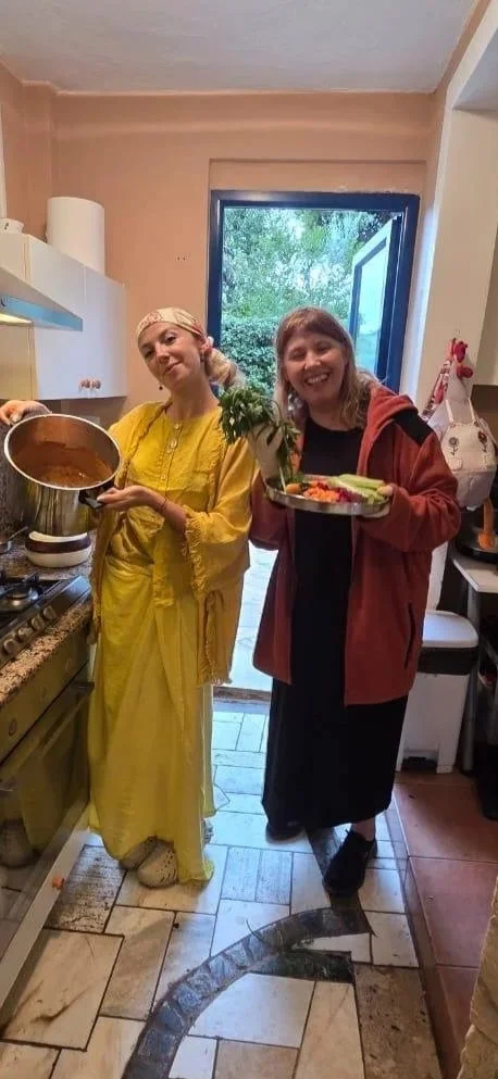 Two women cooking and serving food in a kitchen, one is dressed in traditional Indian attire, the other in casual clothes, both smiling.
