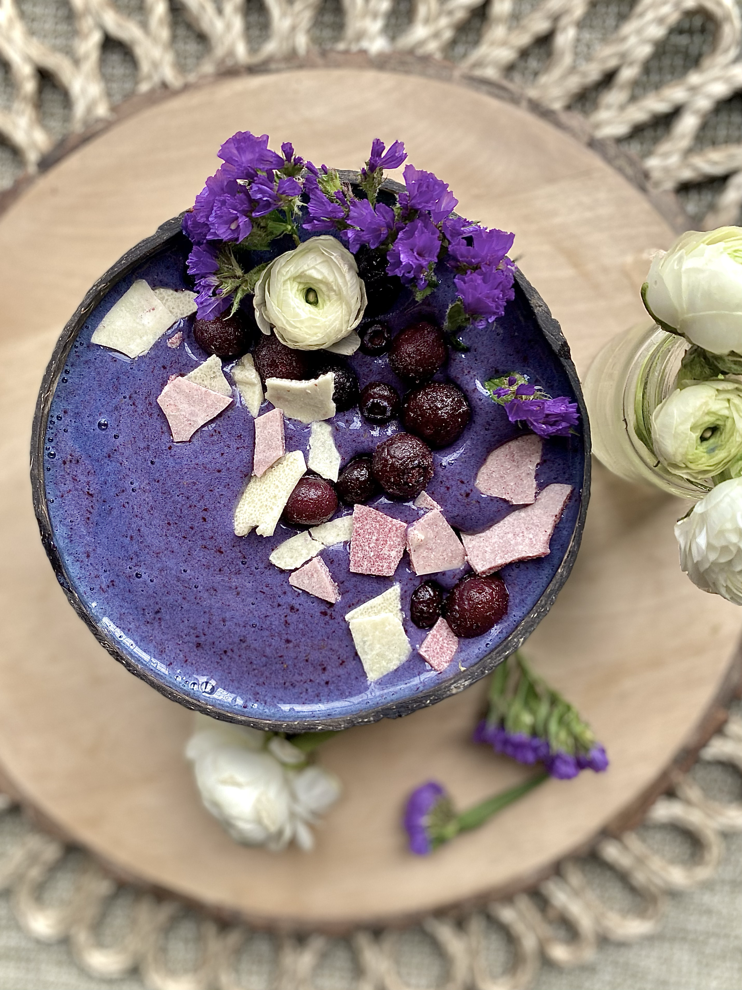 A purple smoothie bowl topped with white, purple, and dark berries, purple flowers, and pink and white coconut flakes on a wooden tray.