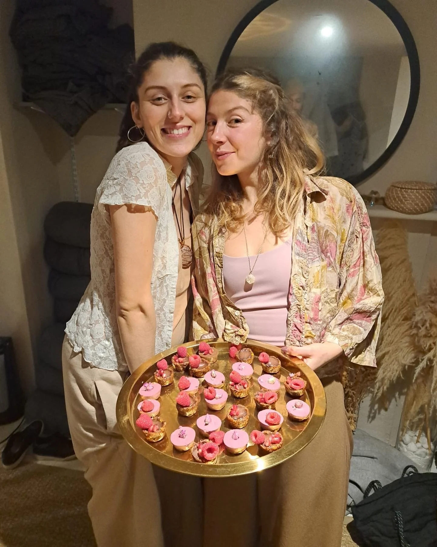 Two women smiling and holding a tray of pink and chocolate desserts with raspberries, in a cozy indoor setting with a round mirror and decorative dried plants in the background.