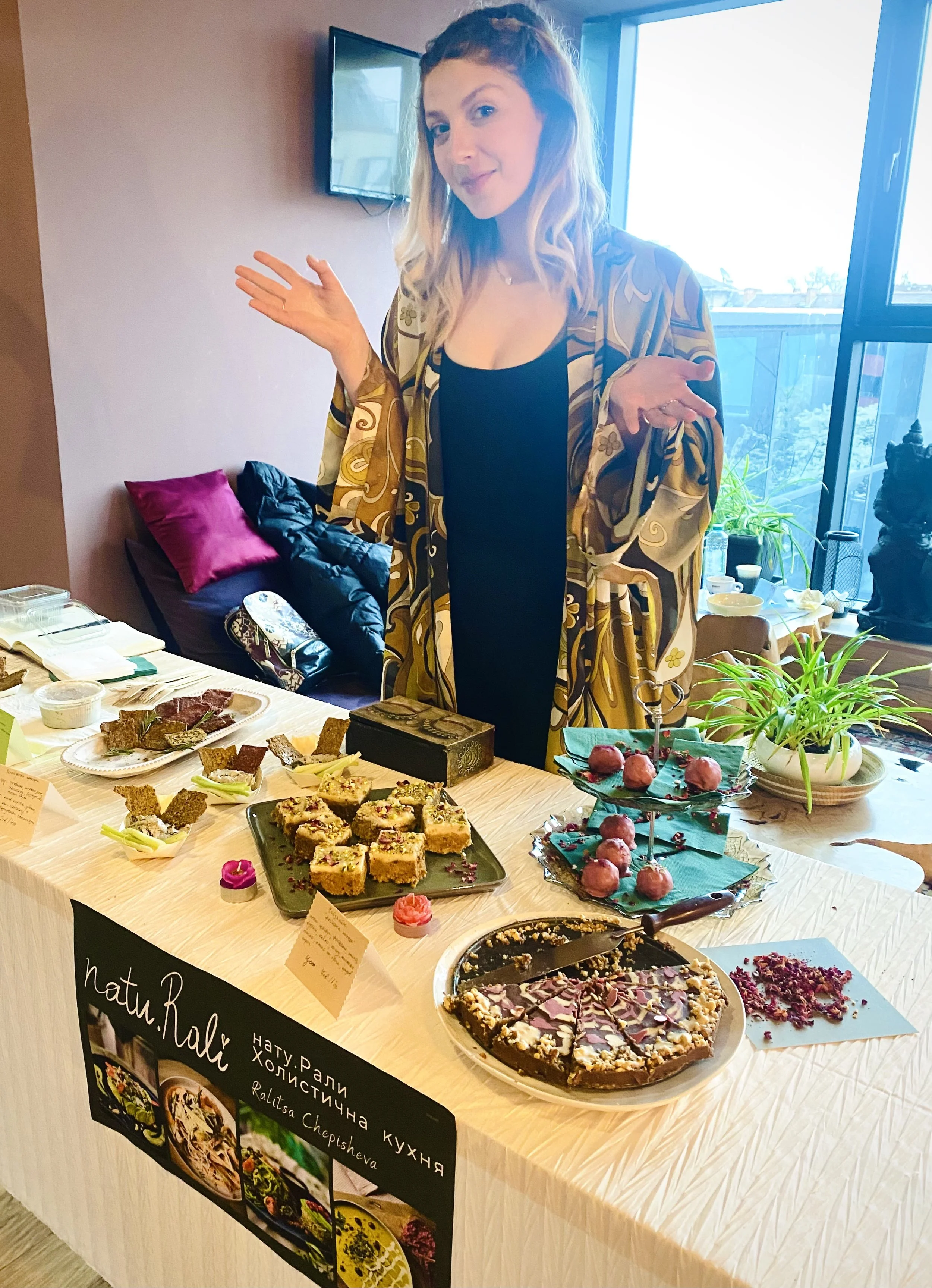 A woman standing behind a table with various desserts and plates, gesturing with her hands, in a room with large windows and a television on the wall.