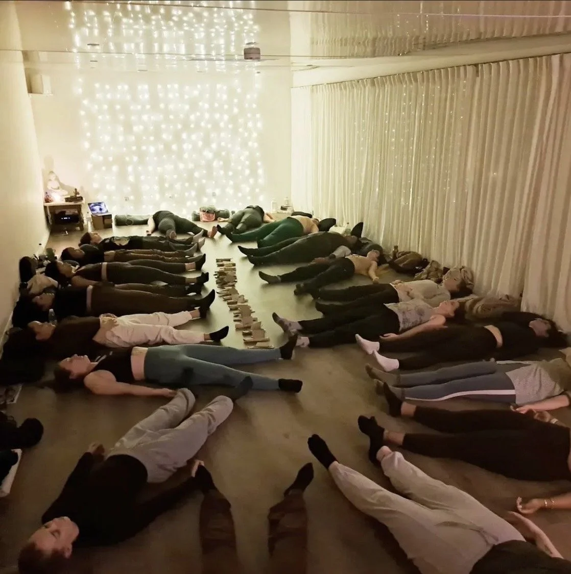 Group of people lying on their backs on the floor with arms at their sides and eyes closed, possibly participating in a relaxation or yoga session, in a room decorated with string lights and curtains.