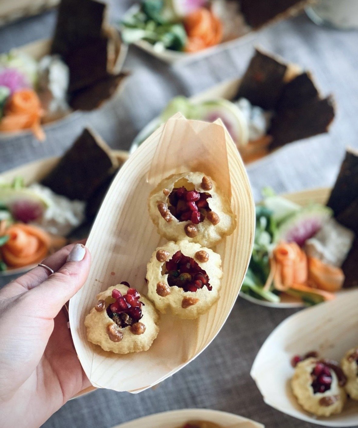 Fried food served on a wooden tray, topped with pomegranate seeds and nuts, held by a person, with other similar dishes in the background.