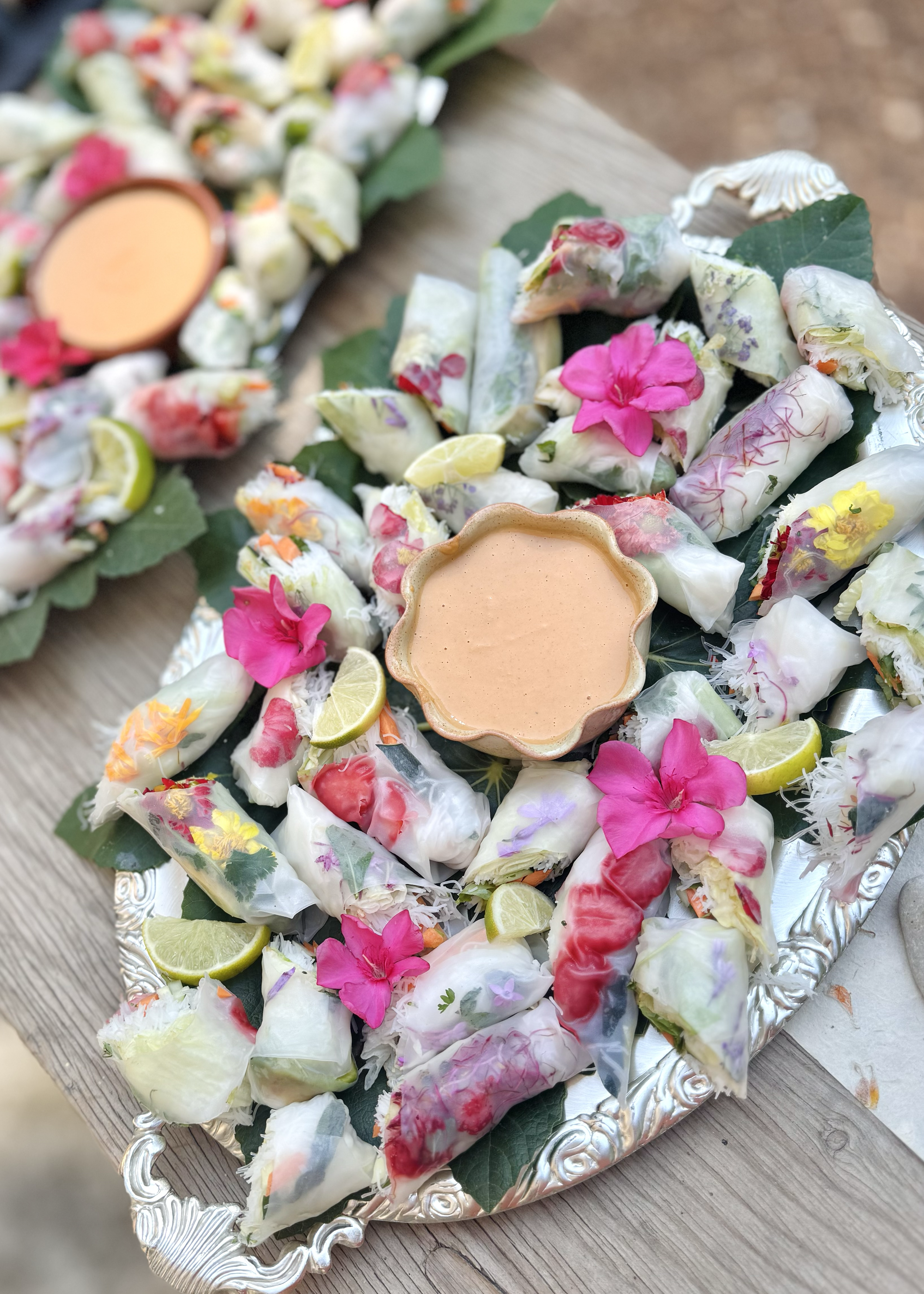 Silver platter with a variety of fresh spring rolls garnished with lime slices, pink flowers, and a bowl of peanut dipping sauce, on a wooden surface.