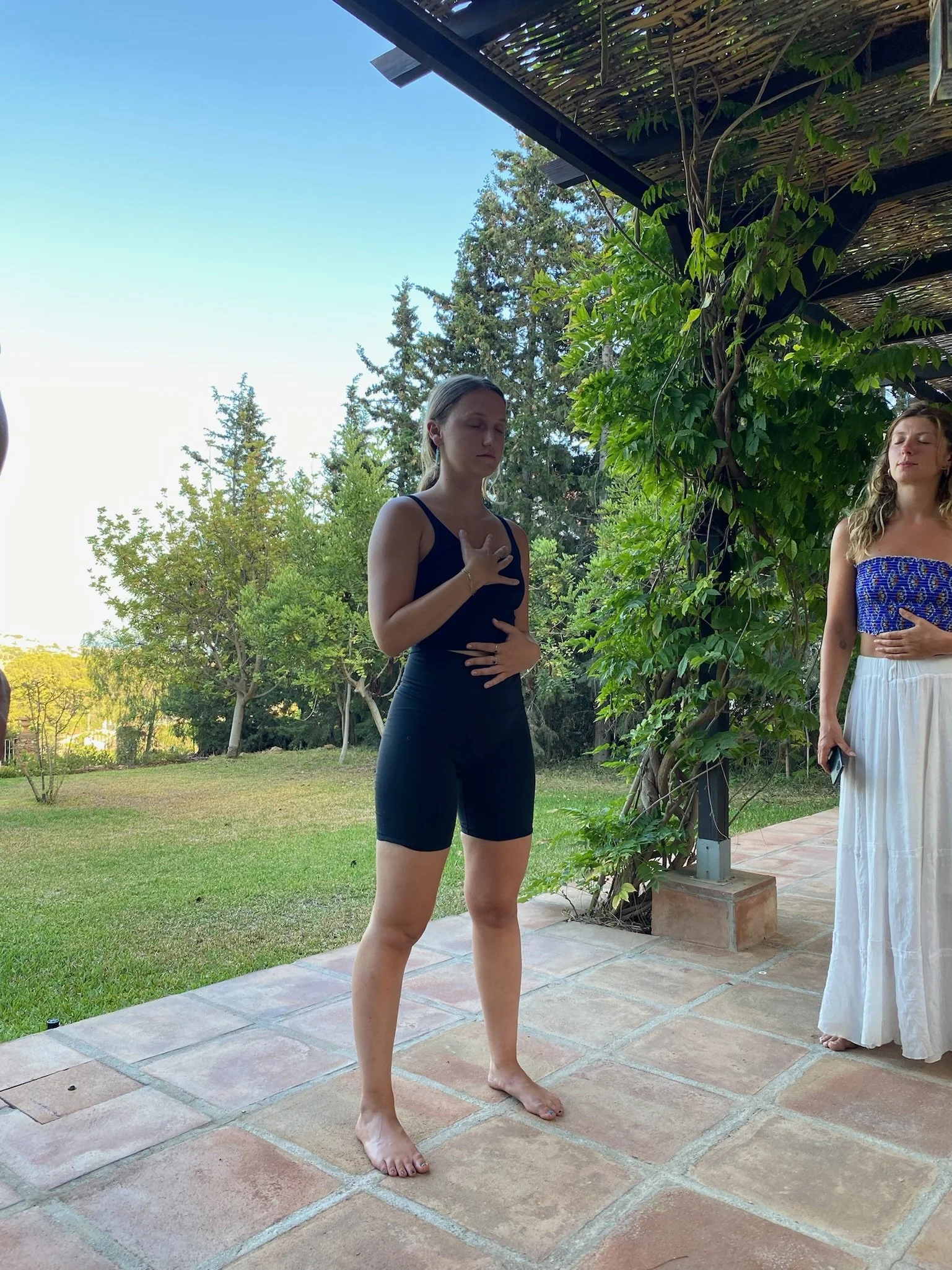 Two women are standing outdoors on a tiled patio, participating in a yoga or meditation practice with closed eyes, one with hand over heart and another with hand on stomach, surrounded by greenery and trees.