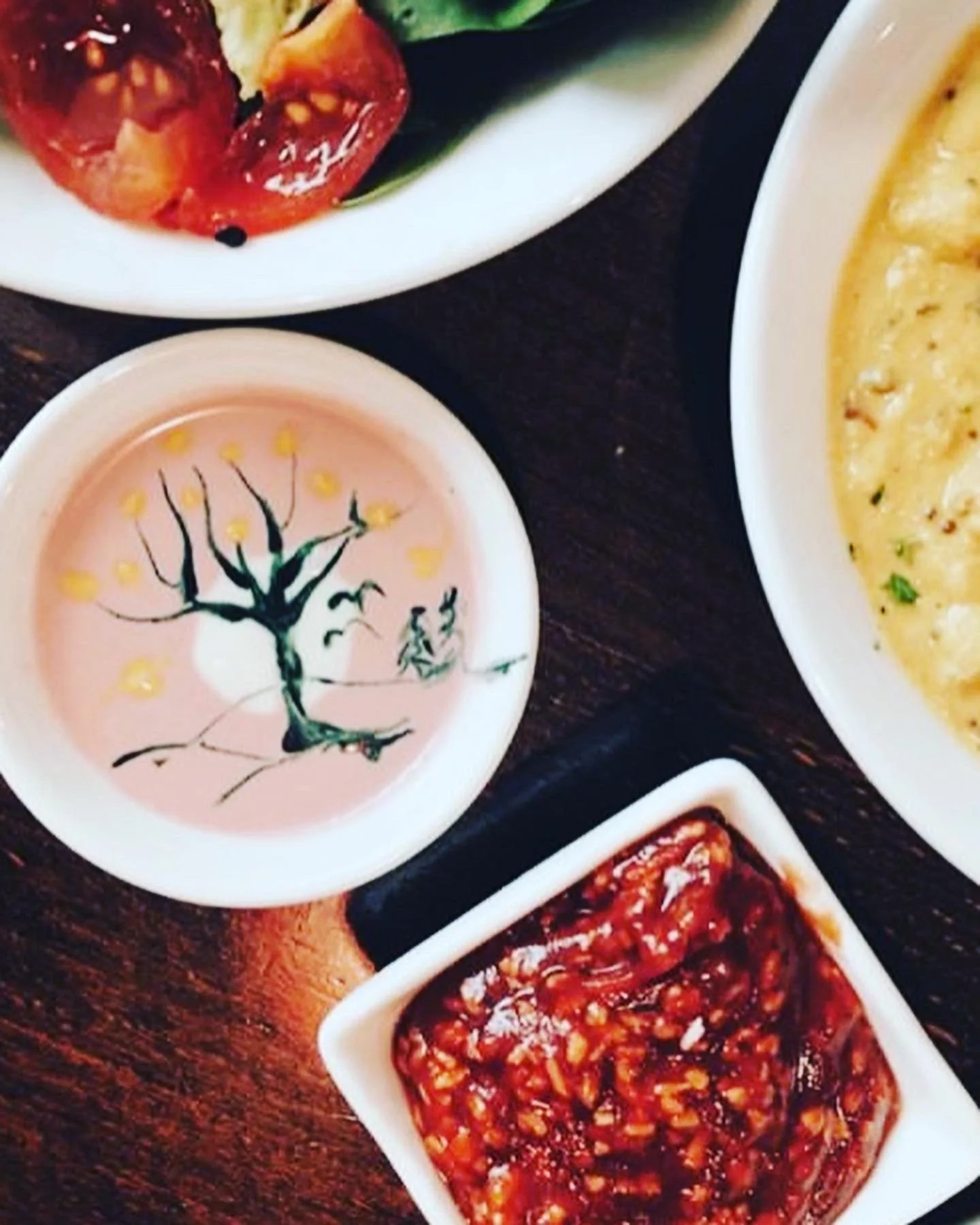 Assorted Korean side dishes including cherry tomatoes, a small dish of pink soy milk with a black tree design, a bowl of thick yellow soup or egg casserole, and a dish of spicy fermented vegetables.