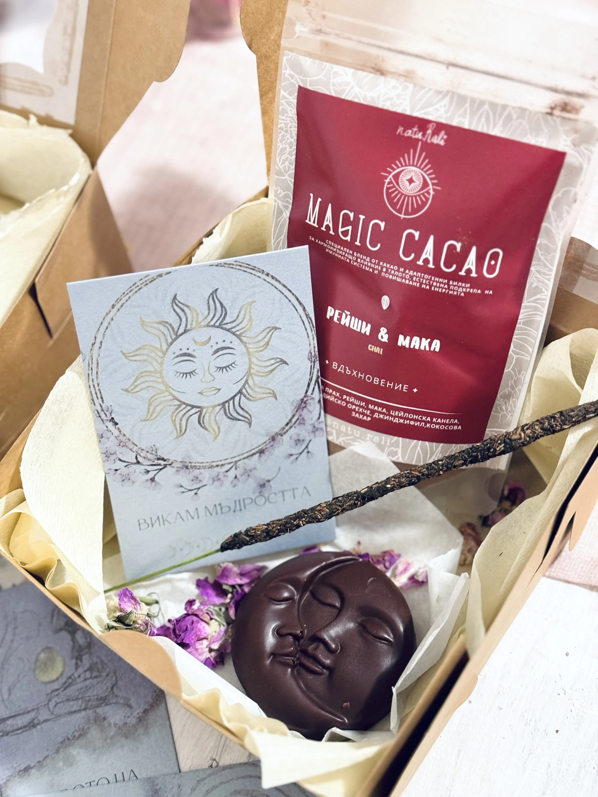 A wooden gift basket contains a massage soap shaped like a face, surrounded by dried purple flower petals, with a red package of magic cacao and a card with an illustration of a sun and moon.