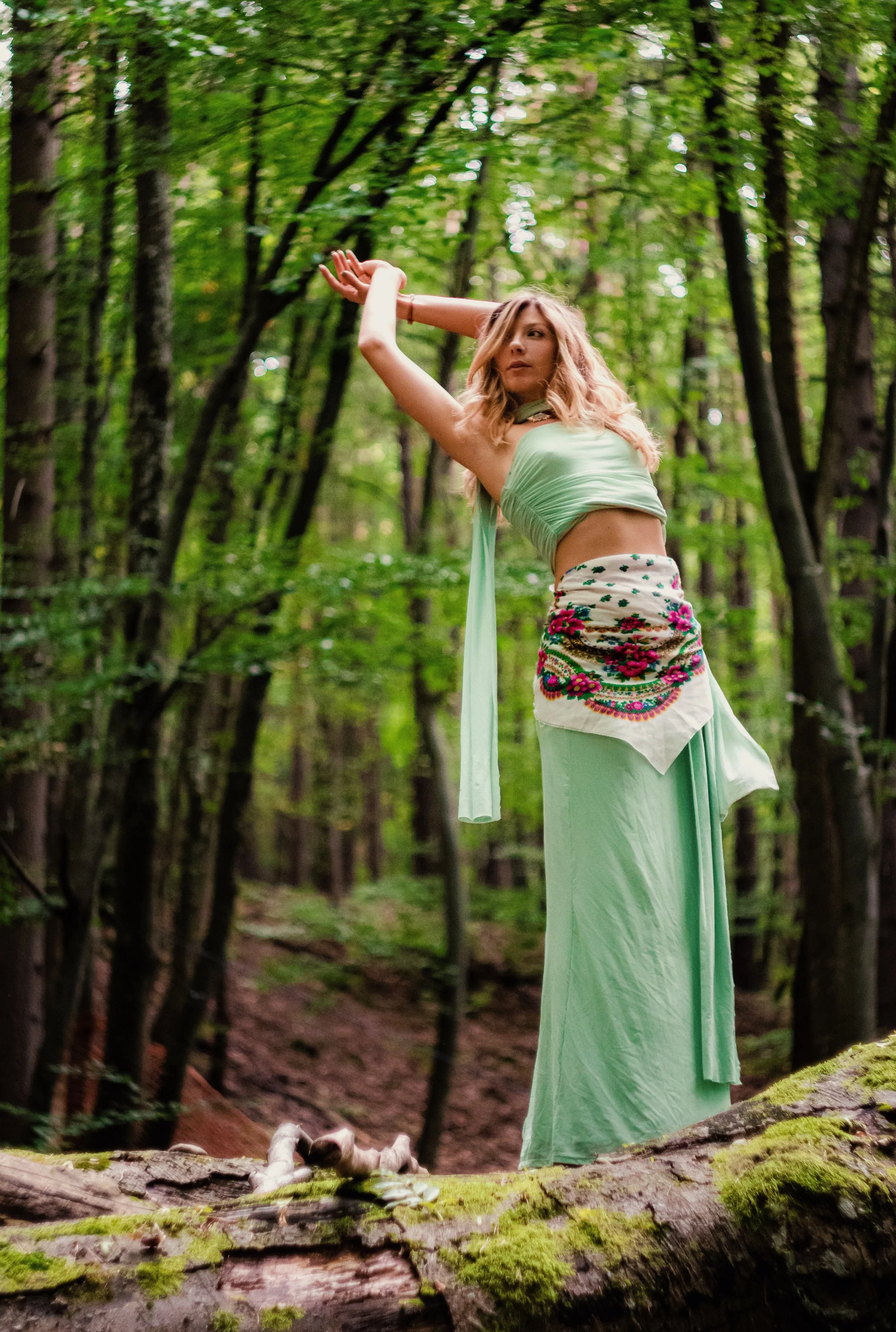 A woman wearing a green wrap top and a long green skirt with floral embroidery poses with her arms raised in a forest. She is surrounded by trees and moss-covered logs.
