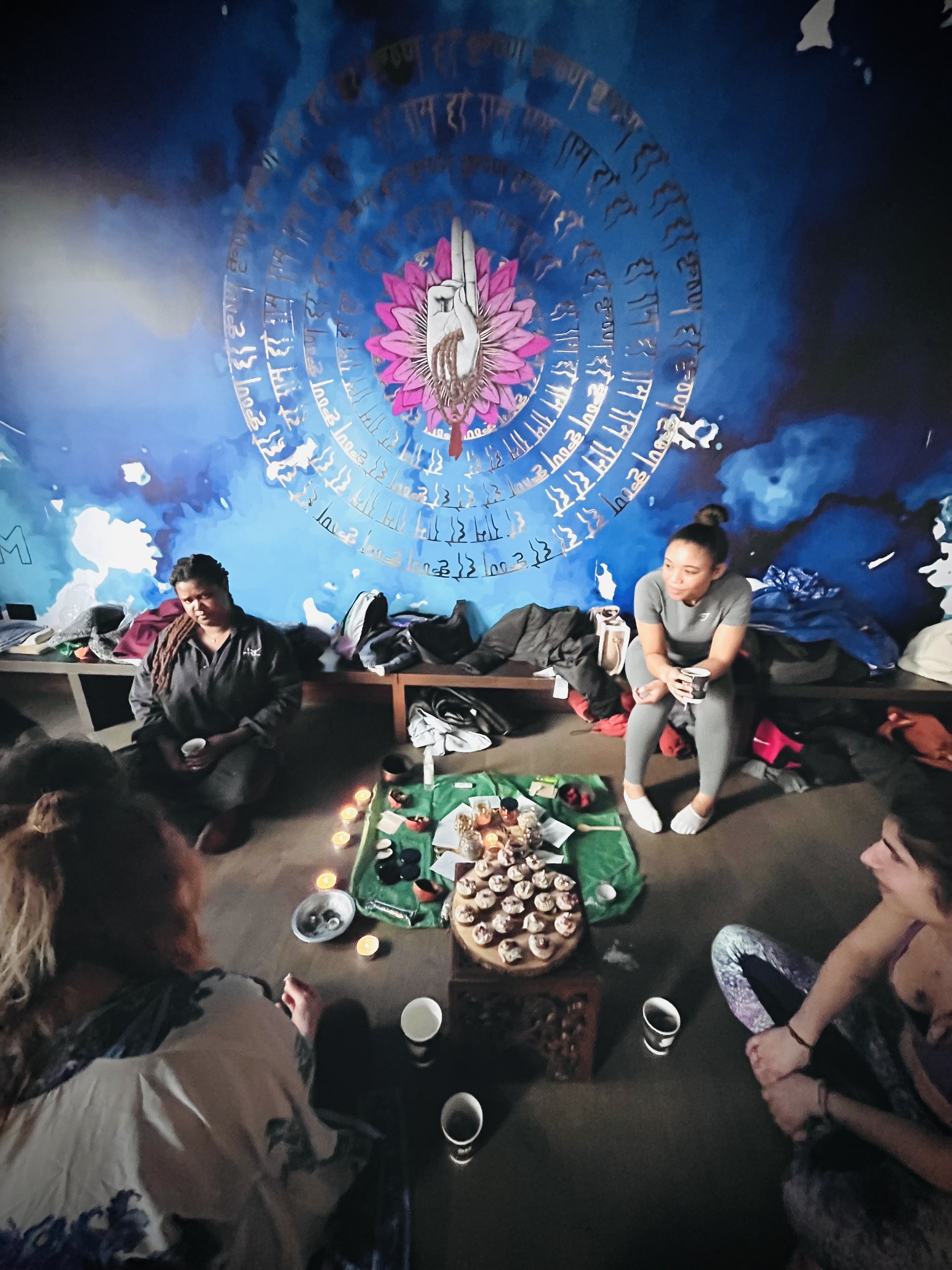 A group of people sitting on the floor around a green cloth with food, drinks, candles, and snacks, in front of a large blue wall with a colorful mural of a hand with a pink lotus and a swan, surrounded by script.