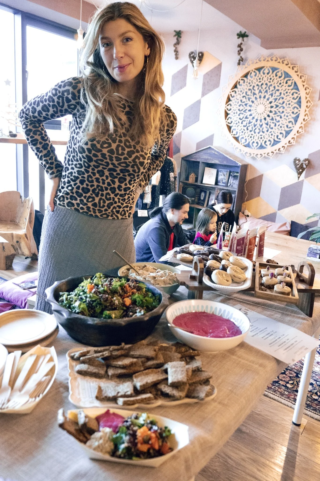 Woman standing near a table with various dishes, including salads, slices of bread, and desserts, during a gathering in a decorated indoor space with people seated in the background.