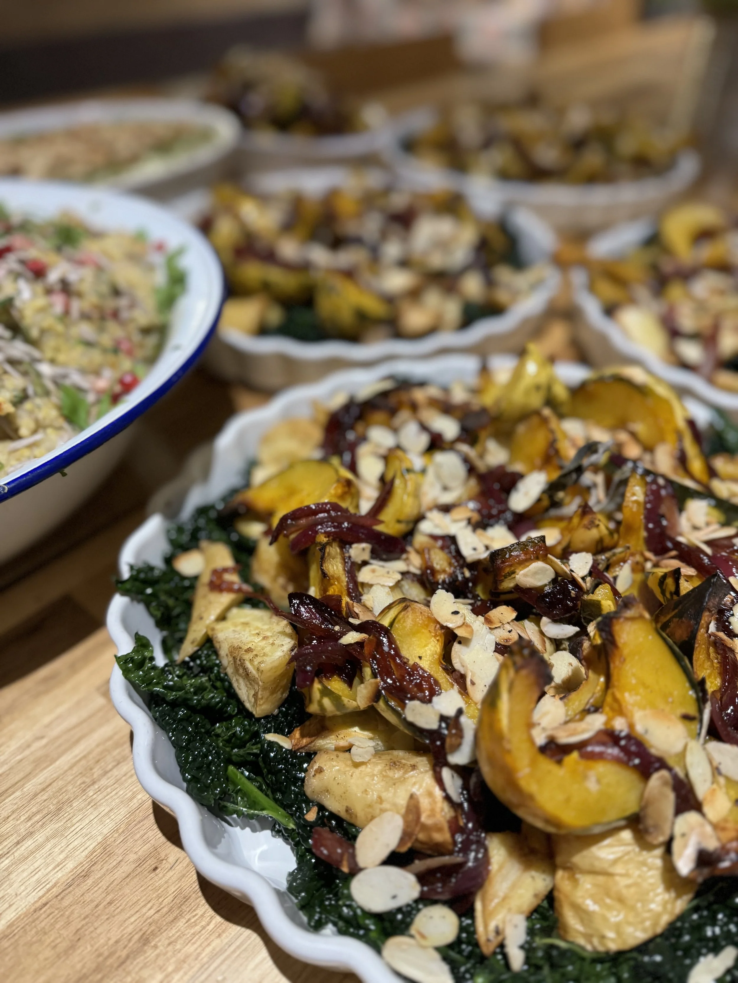 A dish of roasted squash and roasted vegetables topped with sliced almonds, served on a white scalloped plate with a bed of leafy greens, with other similar dishes in the background.