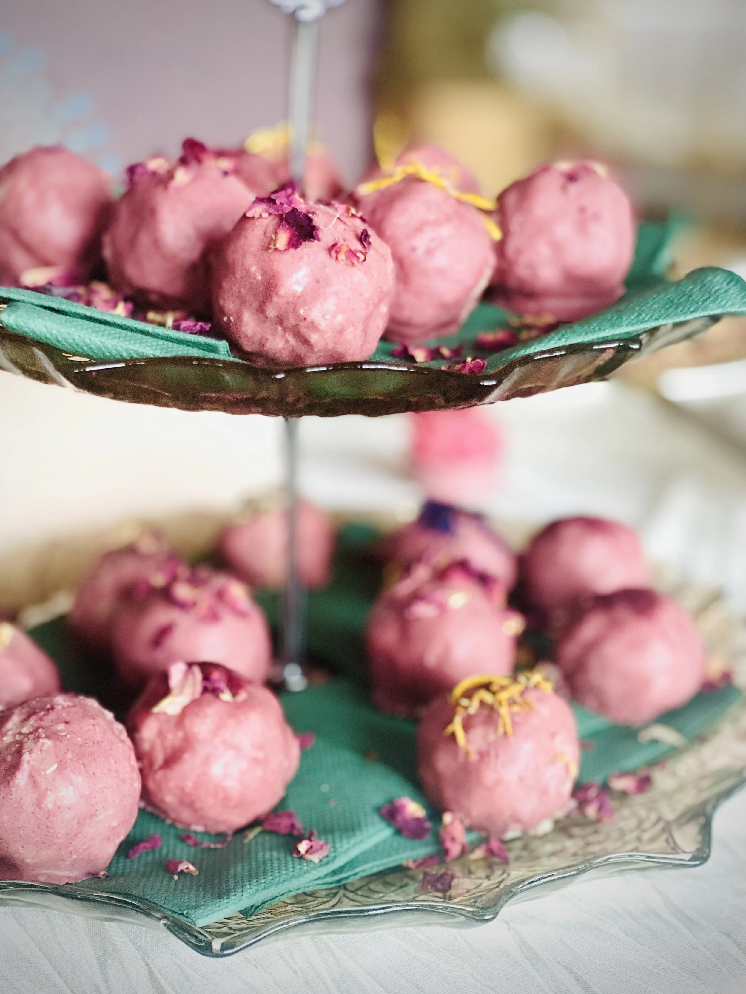 Pink cake pops decorated with pink and yellow flower petals on a glass tiered stand with a green napkin lining each layer.