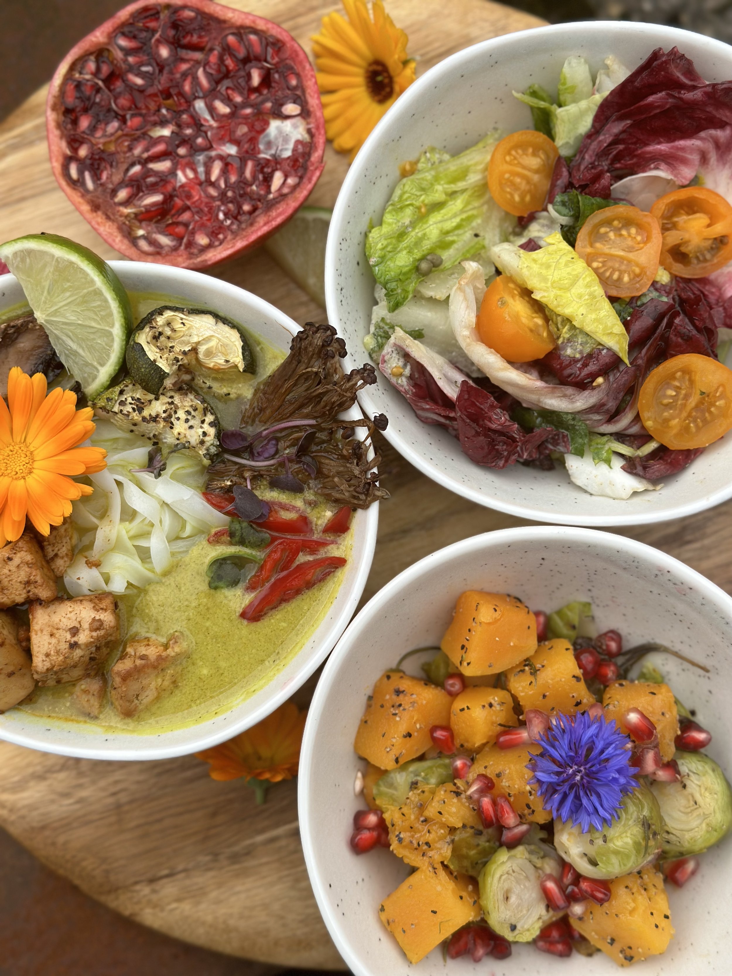 Three bowls of colorful salads with flowers and a halved pomegranate on a wooden surface.