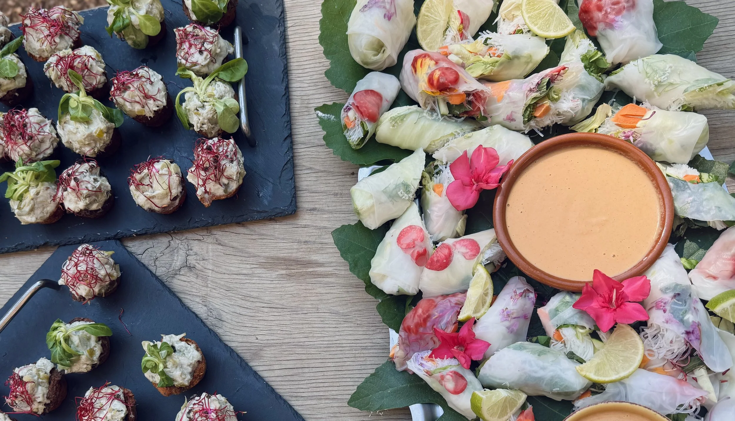 Various appetizers including mini tostadas topped with beans, cream, and microgreens on the left, and a platter of fresh spring rolls with vegetables and herbs, alongside a bowl of dipping sauce on the right, all on a rustic wooden table.
