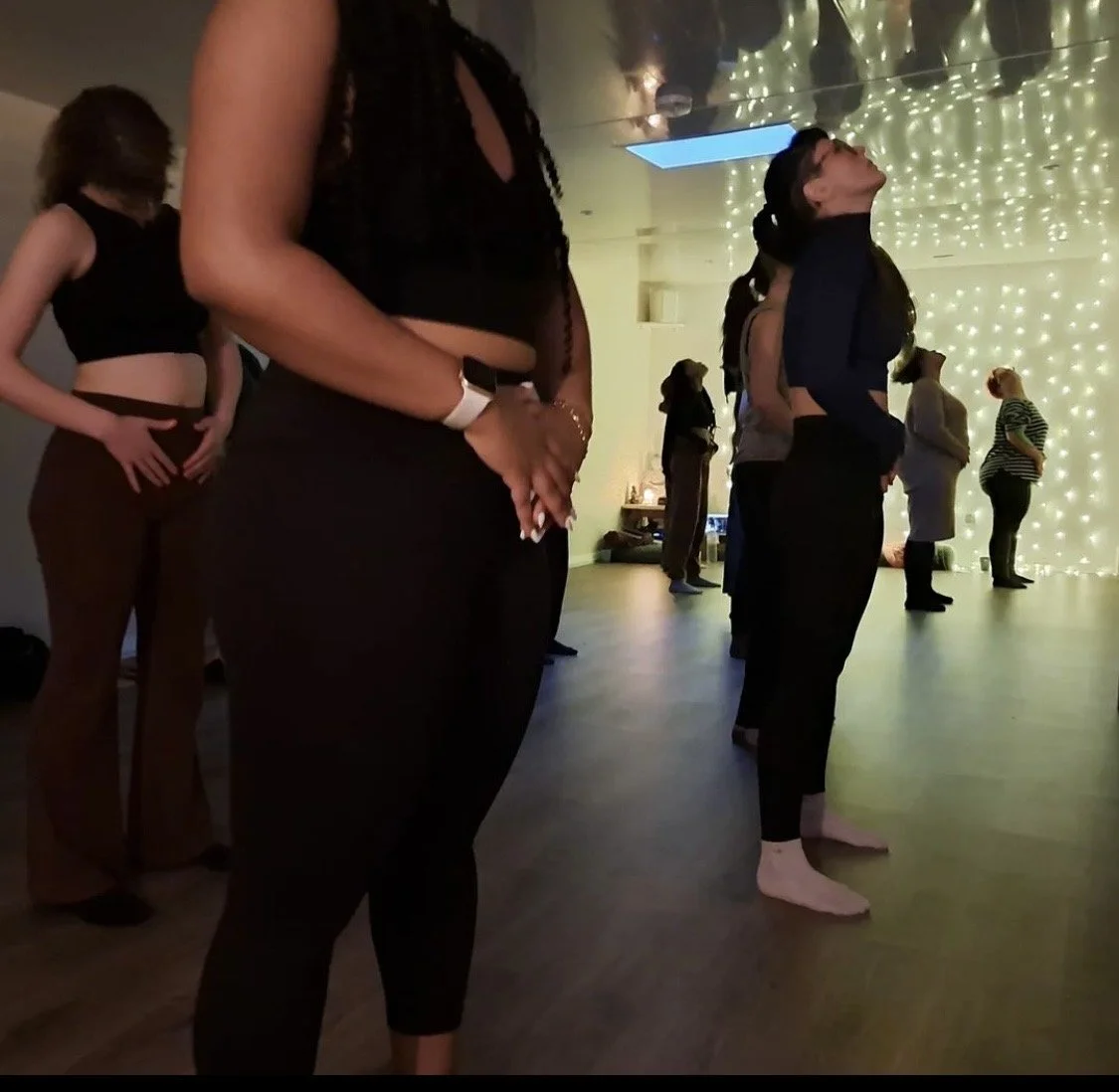 People participating in a meditation or yoga class in a dimly lit room with string lights on the wall, with some standing and some with eyes closed, in various poses and postures.
