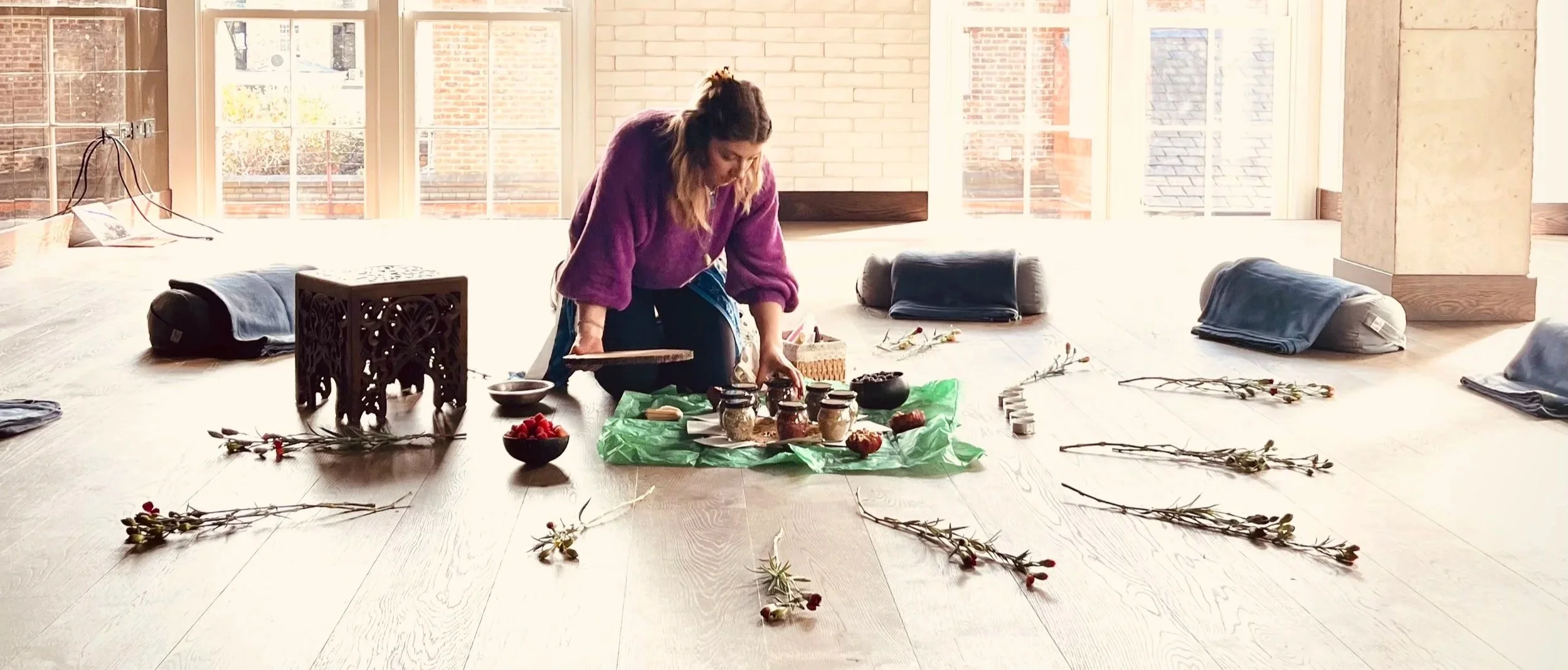 Woman arranging flower stems and jars on green paper in a room with large windows, cushions on the floor, and natural light.
