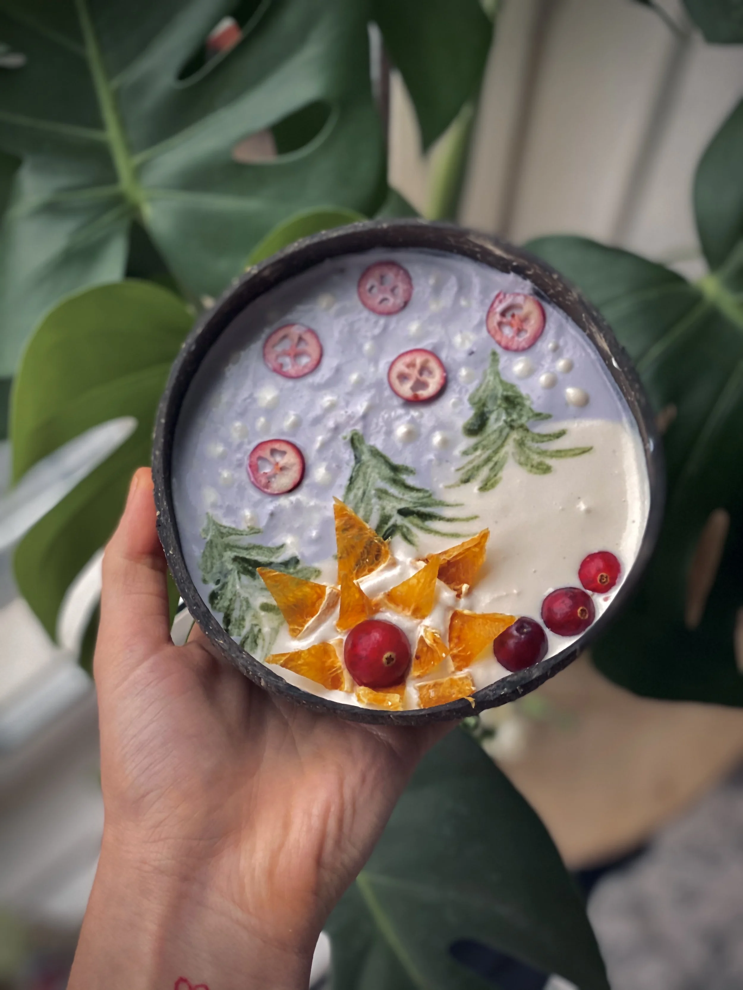 A bowl of creamy dessert decorated with slices of chili peppers, orange pieces, red berries, and green herb leaves, set against large green plant leaves.
