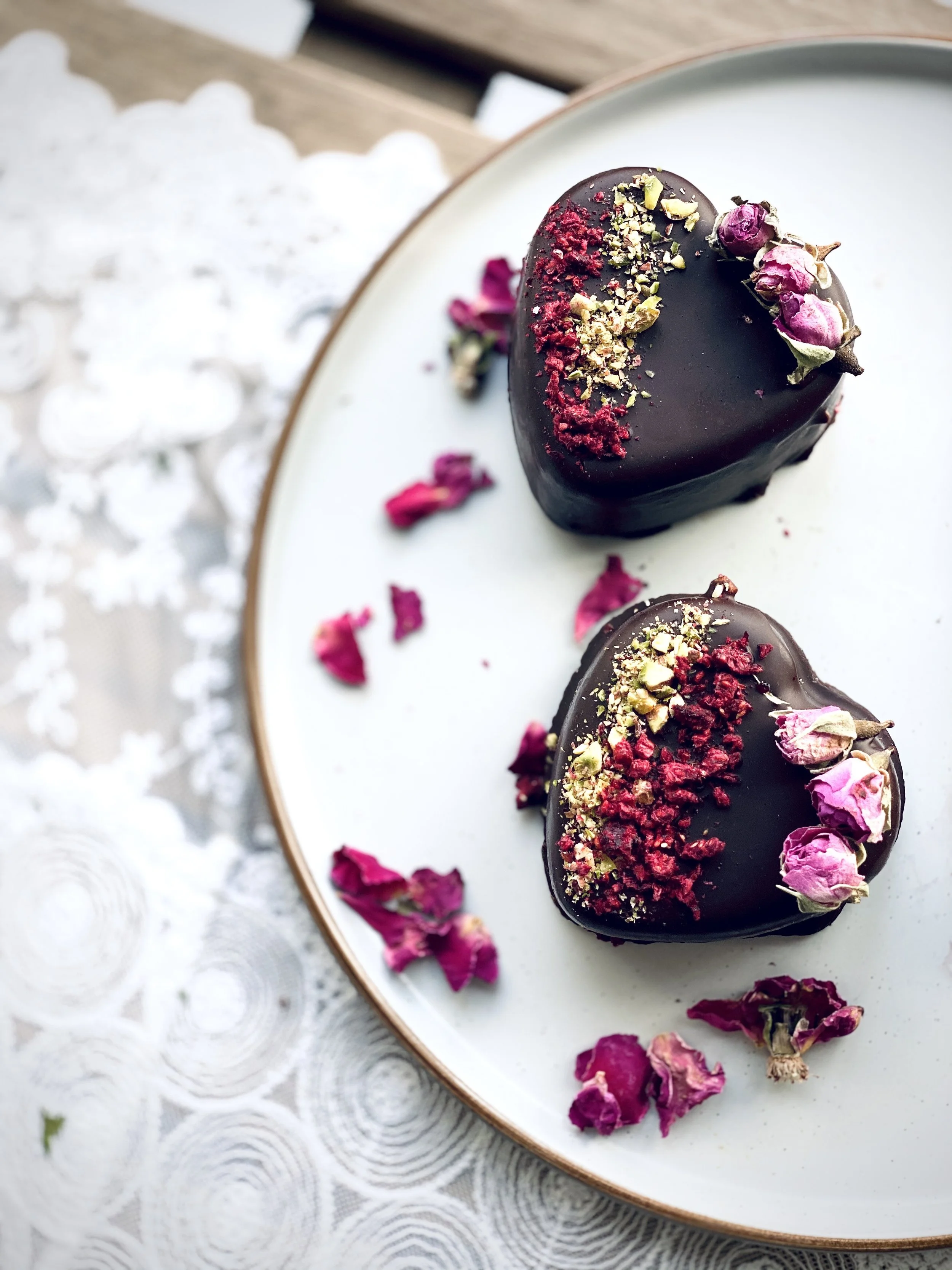 Two heart-shaped chocolates coated in dark chocolate with pink dried rosebuds and crushed pink and green toppings, garnished with scattered dried rose petals on a white plate.