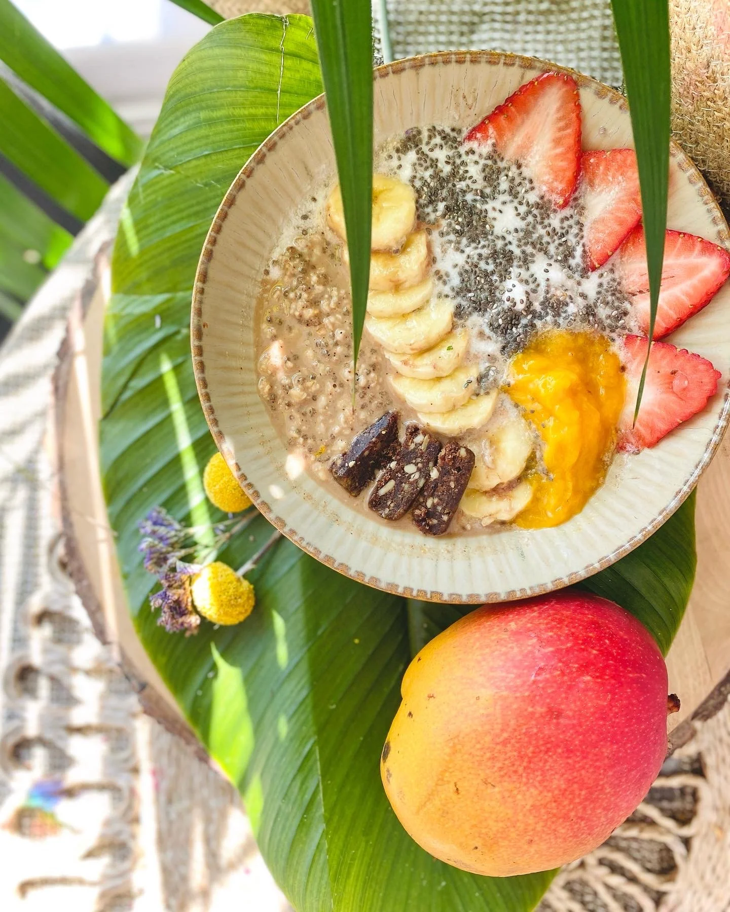 A bowl of fruit and chia seed pudding placed on a large green leaf, with an apple beside it, and tropical plants and flowers in the background.