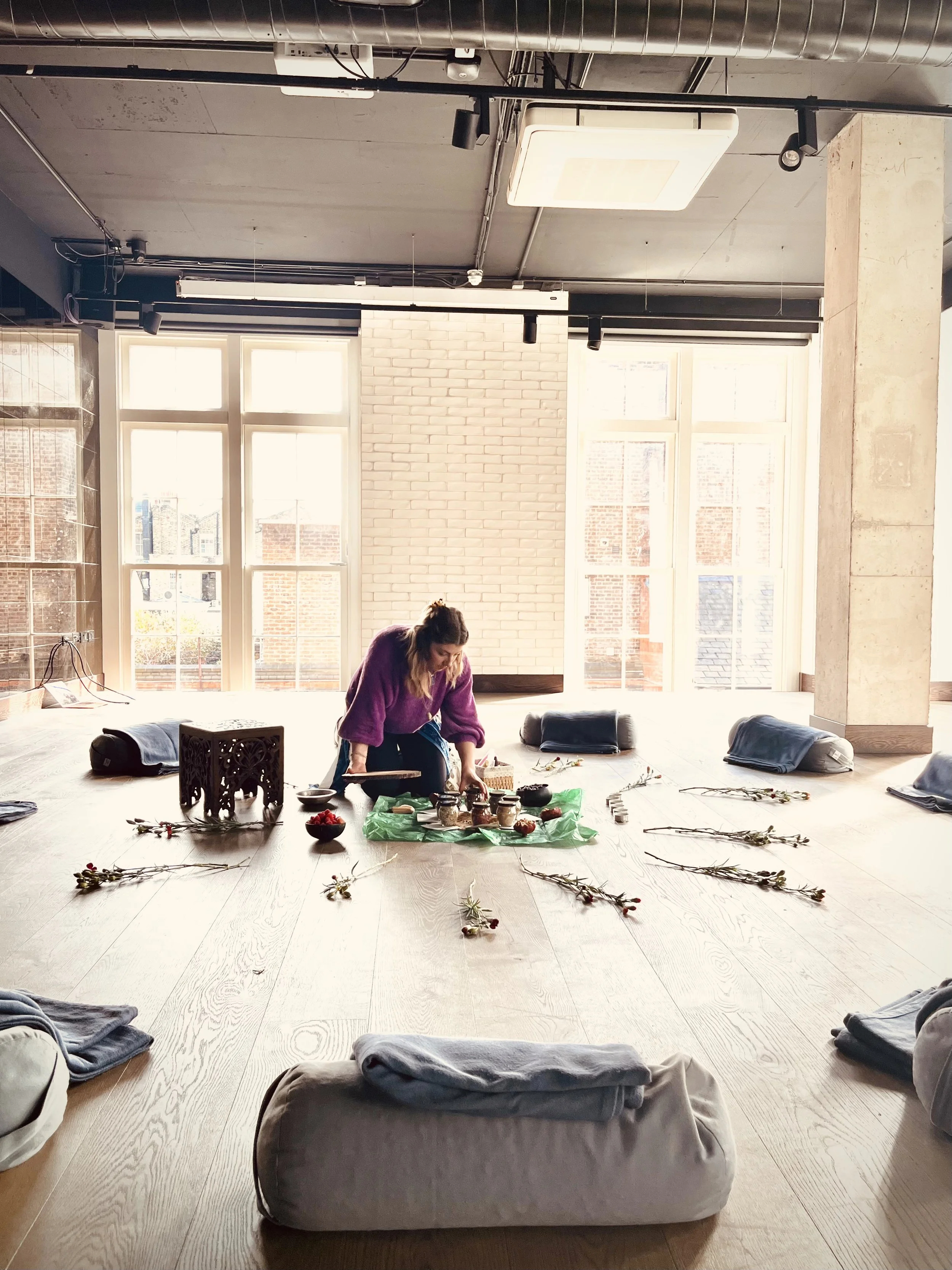 A woman in a purple sweater arranging items on a green cloth on the floor. The room has large windows, pillows on the floor, and dried flowers scattered around for what appears to be a spiritual or meditation session.