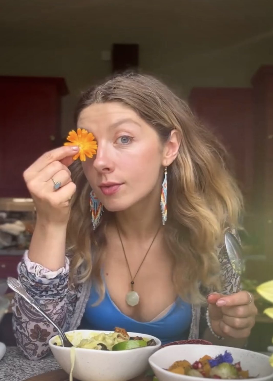 Woman with wavy hair holding a yellow flower near her eye, sitting at a table with bowls of food, wearing colorful earrings and a patterned top.
