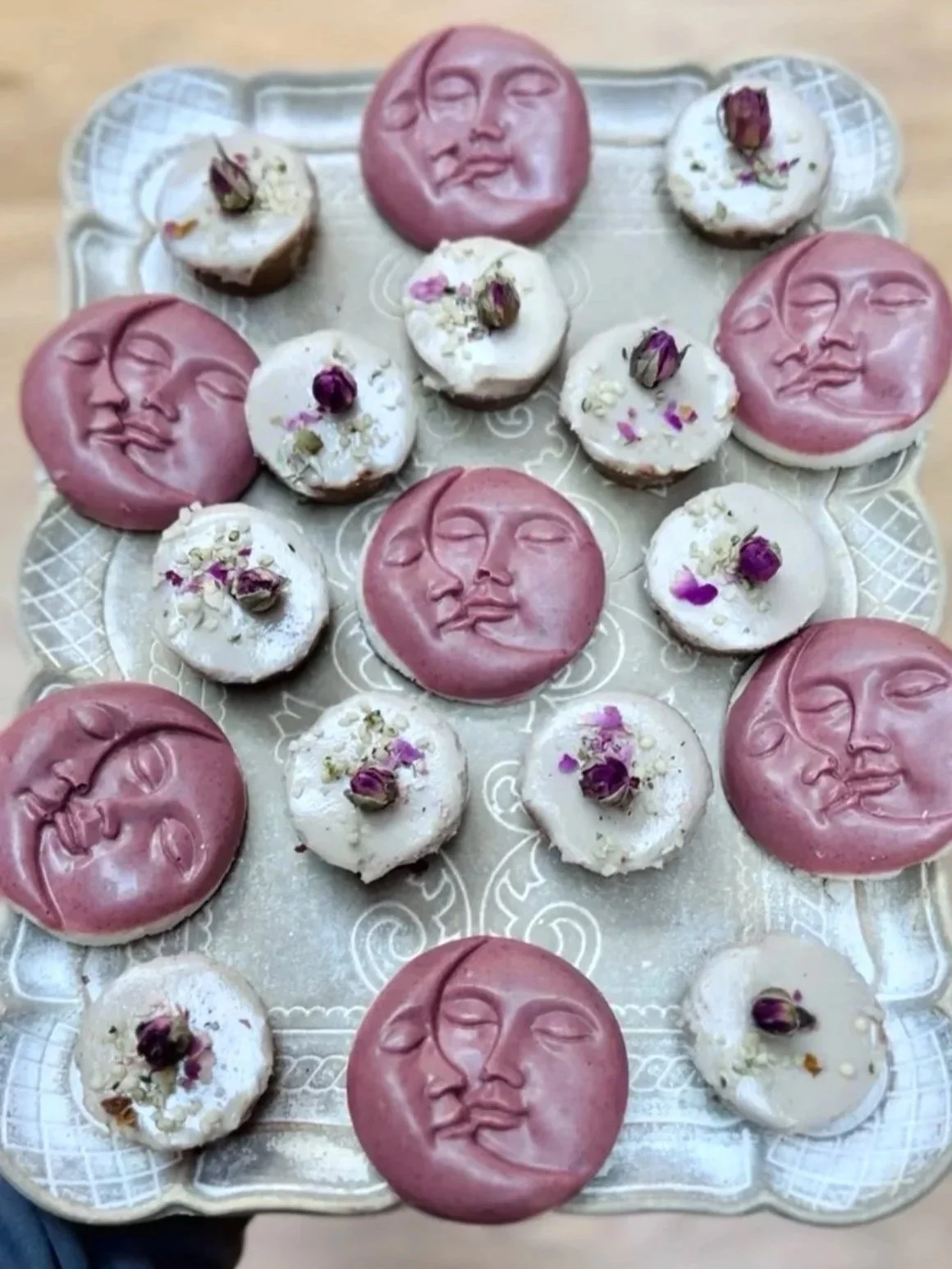 Assorted cookies with moon and face designs, topped with dried rose petals, arranged on a silver tray.