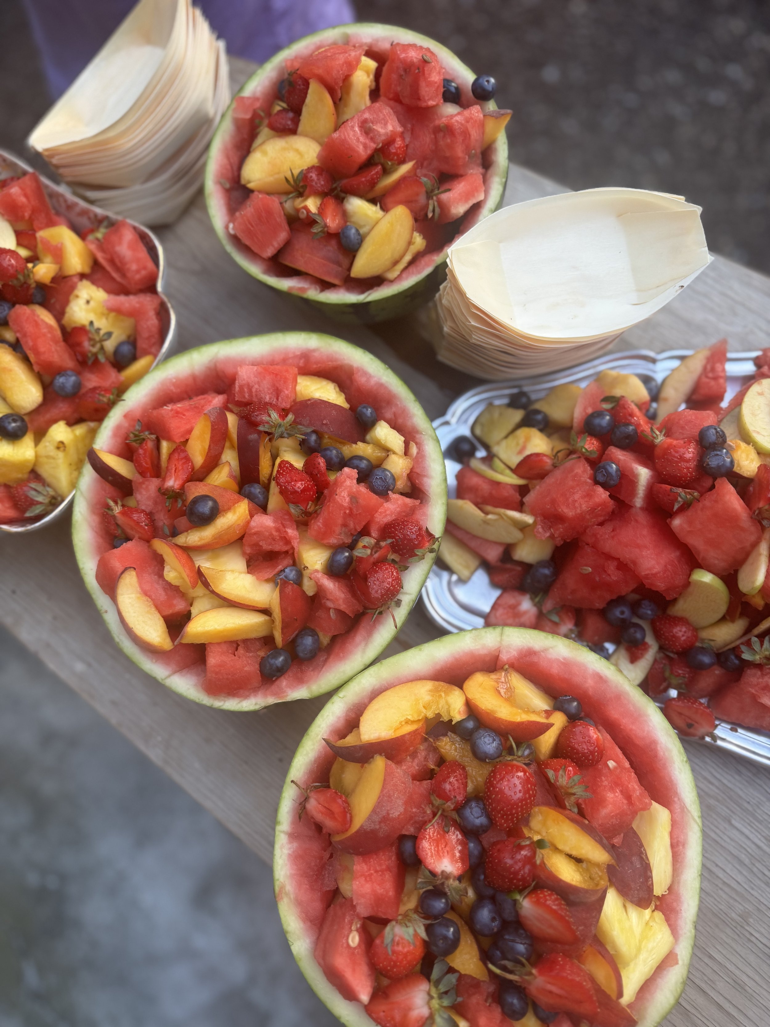 Watermelon halves filled with mixed fruit including strawberries, blueberries, watermelon pieces, peaches, and other seasonal fruits displayed on a wooden table.