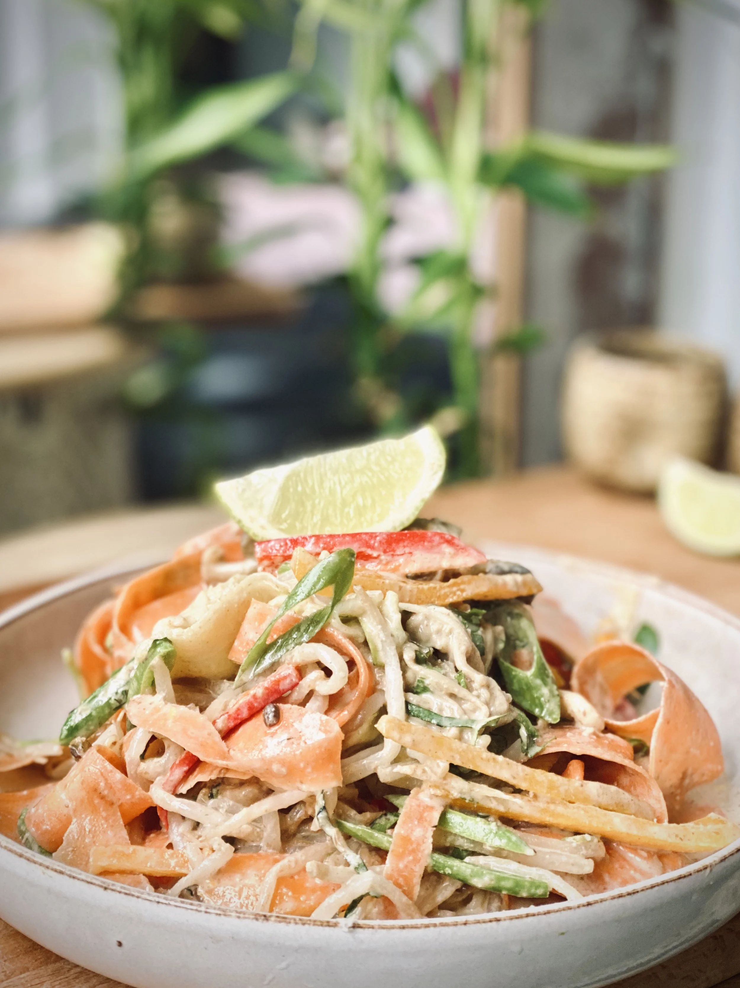 Close-up of a plate of colorful vegetable pasta salad garnished with a lime wedge, placed on a wooden table with blurred indoor plants and decor in the background.