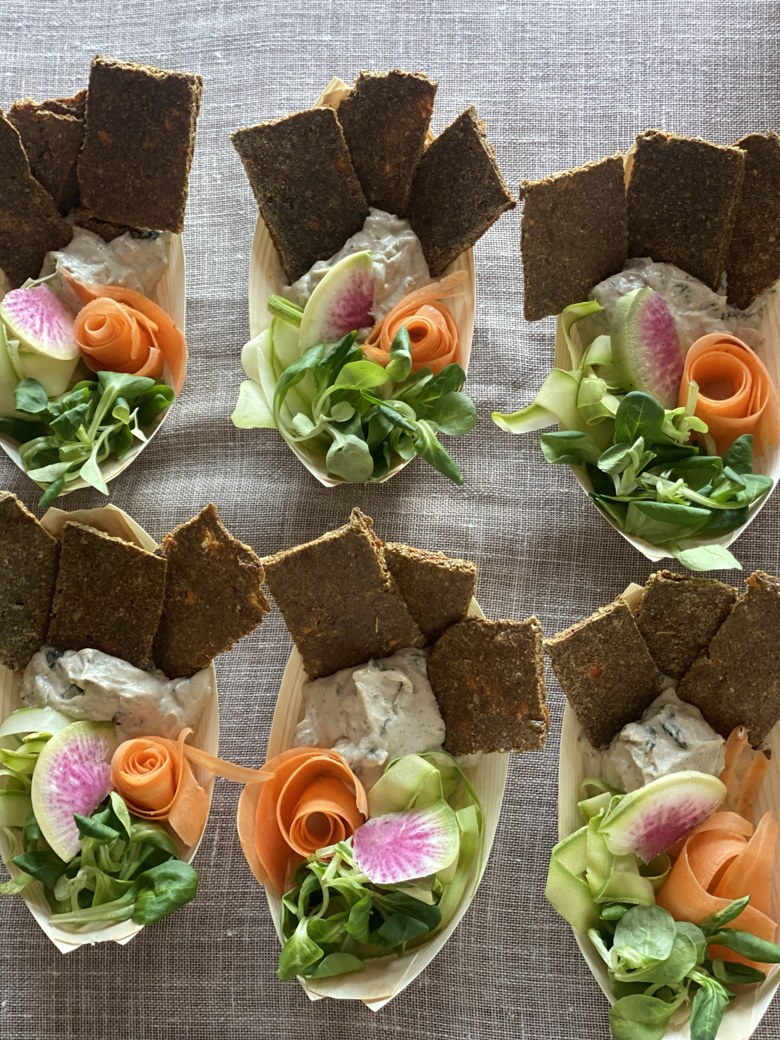 Six small appetizer bowls with greens, sliced vegetables, and crispy bread slices on top, placed on a textured beige surface.
