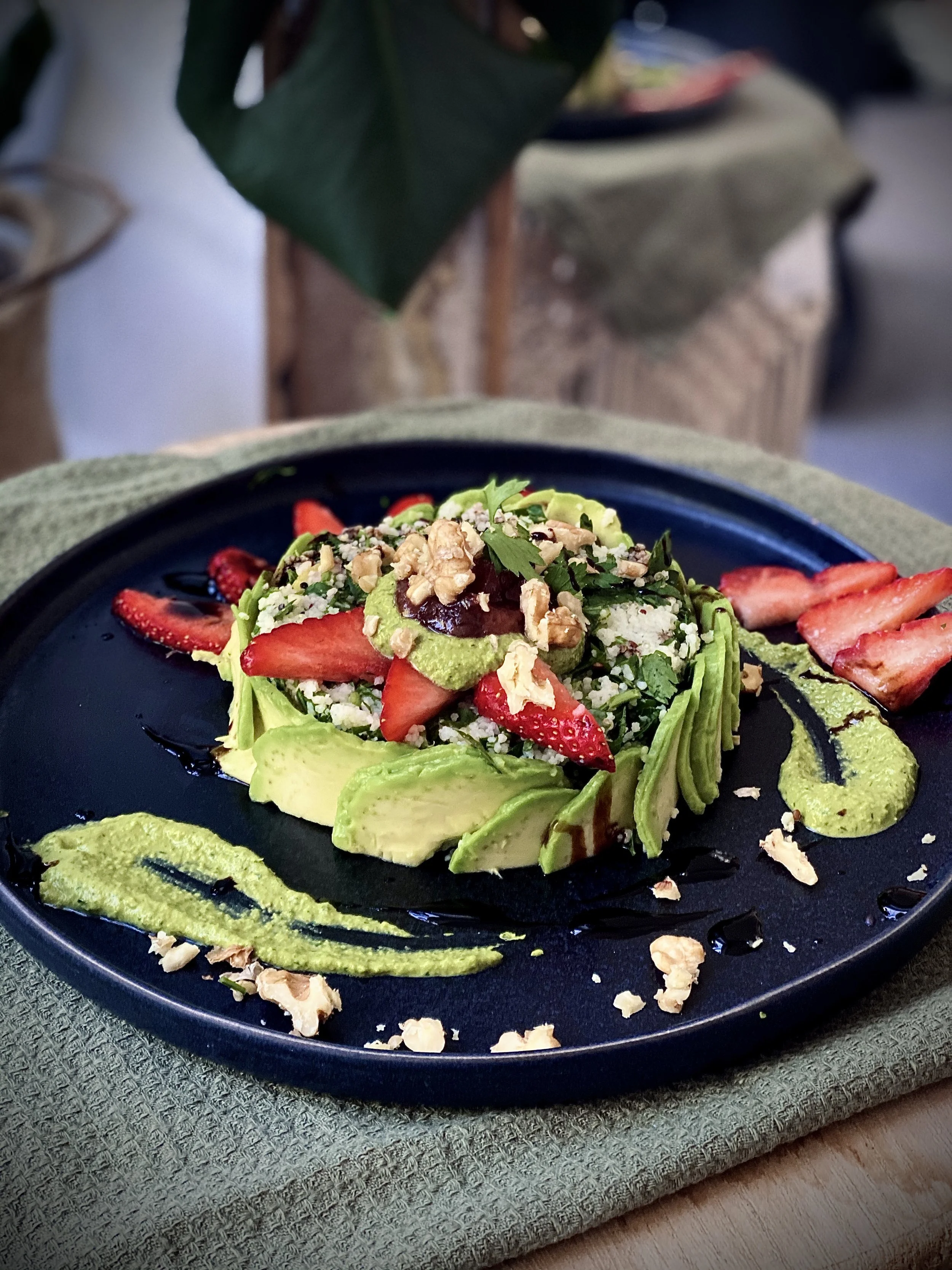 A dark plate of avocado salad garnished with strawberries, nuts, and drizzled with balsamic glaze, served with a swirl of green sauce and crumbled cheese, on a textured mat.