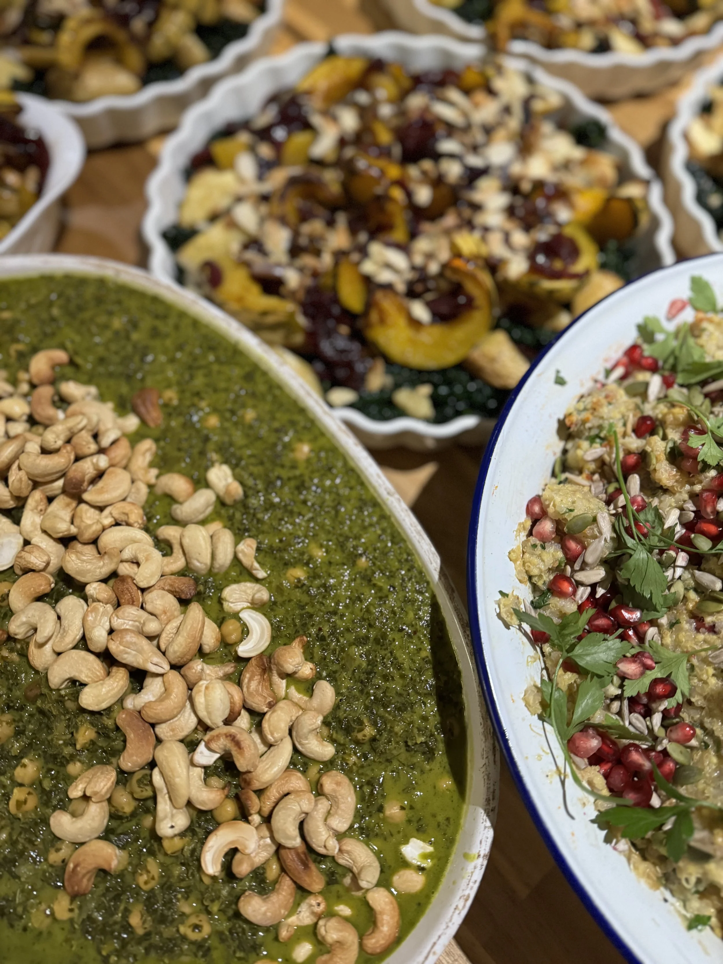 Assorted dishes including a green sauce topped with cashews, a baked dish with yellow squash and cranberry topping, and a grain salad with pomegranate seeds and herbs.