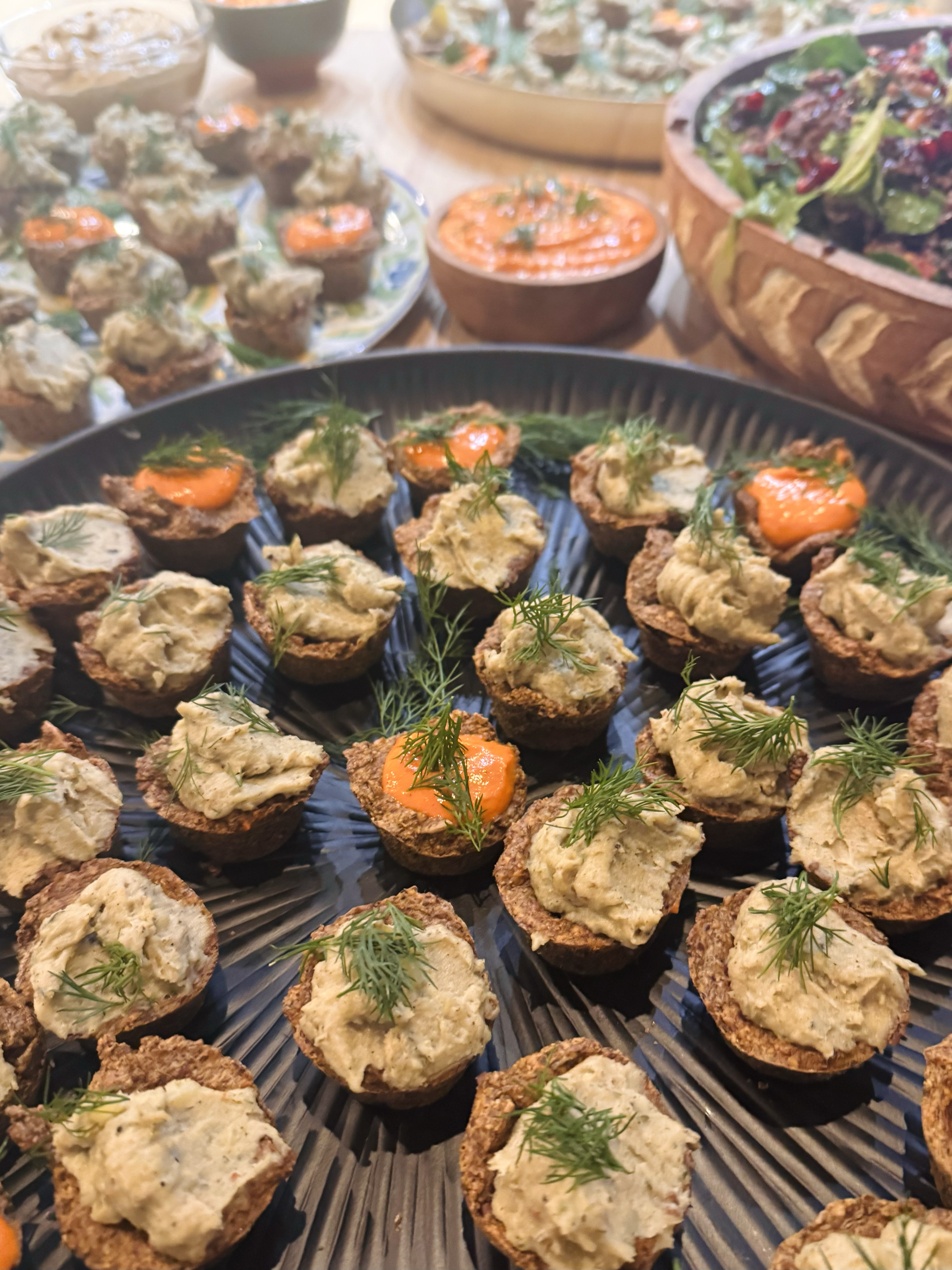 A platter of bite-sized appetizers topped with a creamy mixture and garnished with fresh dill, with other dishes and bowls of food in the background.