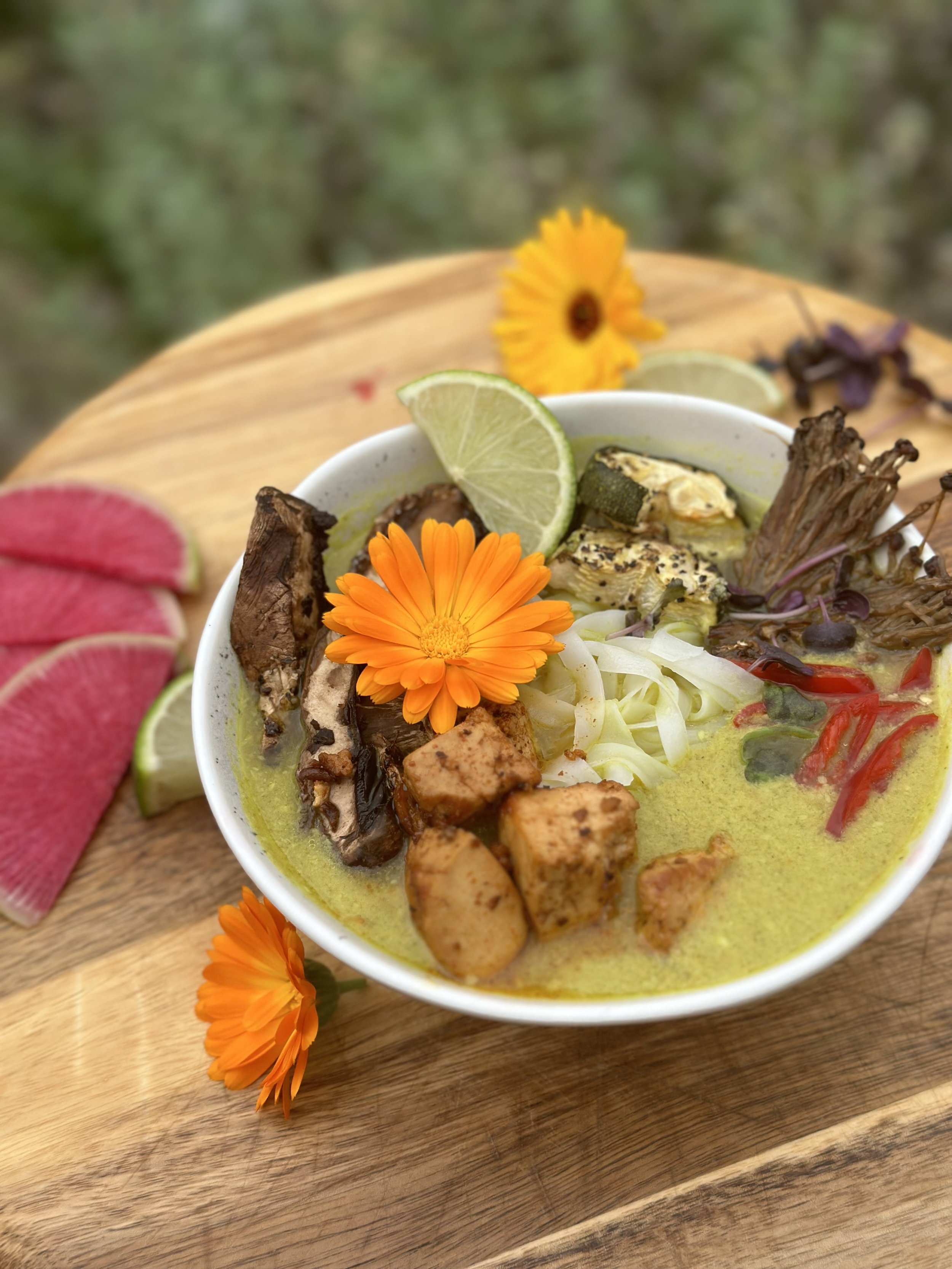 A bowl of vegan curry garnished with edible flowers, lime slices, and vegetables on a wooden board, with watermelon slices and flowers in the background.