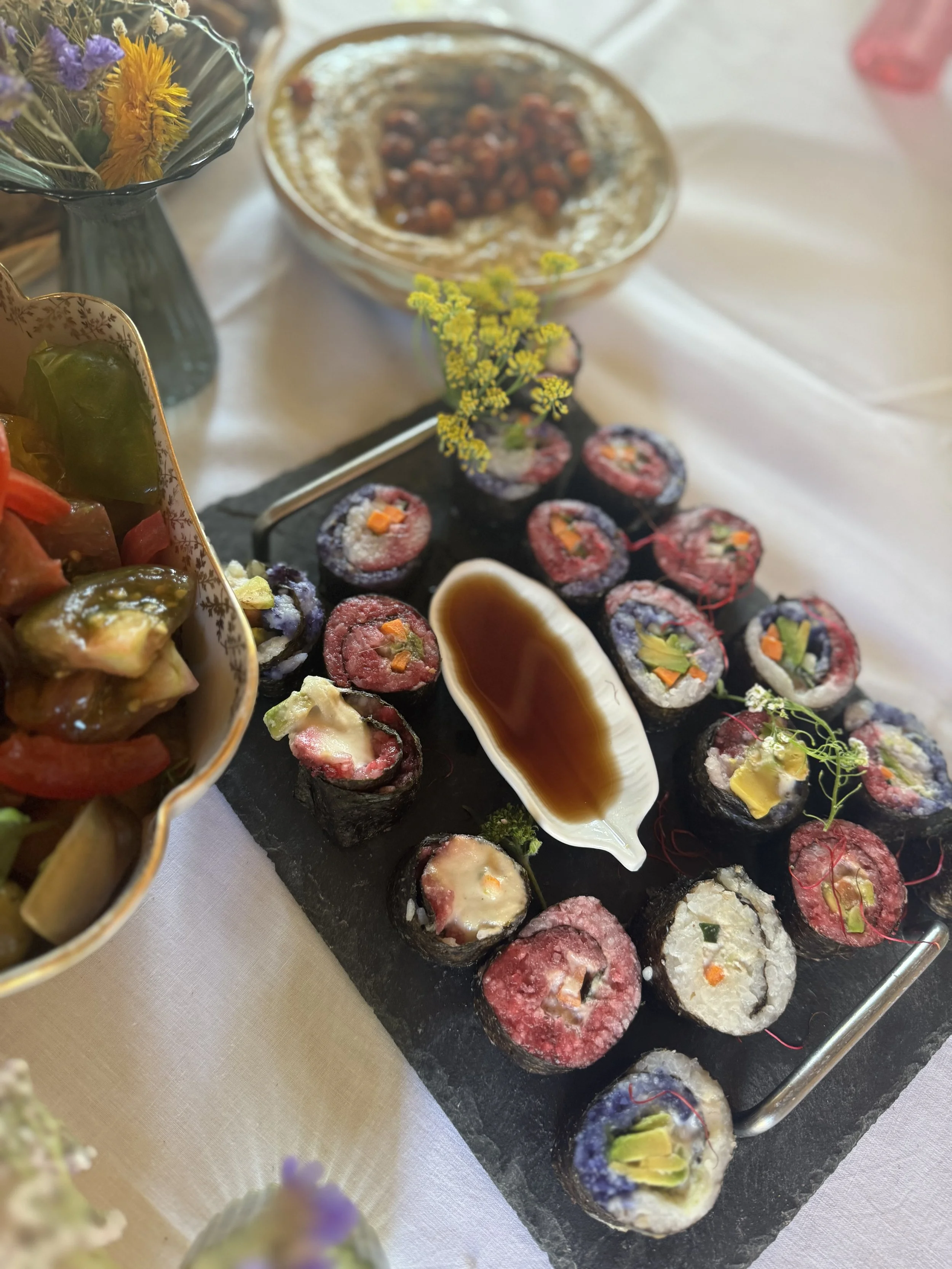 Assorted colorful sushi rolls on a black slate platter with soy sauce in a shell-shaped dish, with other dishes and flowers nearby.