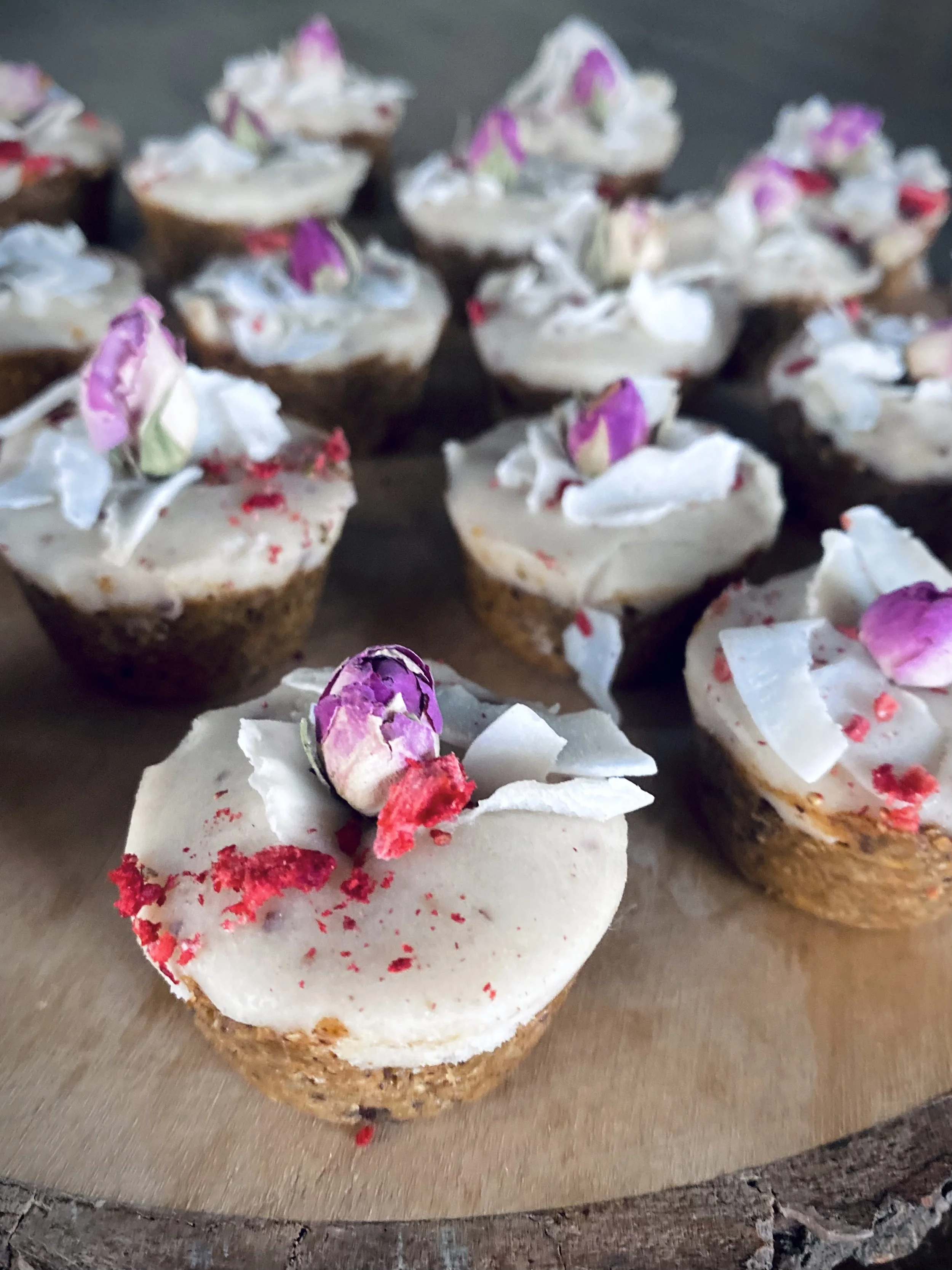 Cupcakes with white frosting, pink and purple flower petals, and red and white sprinkles on a wooden surface.