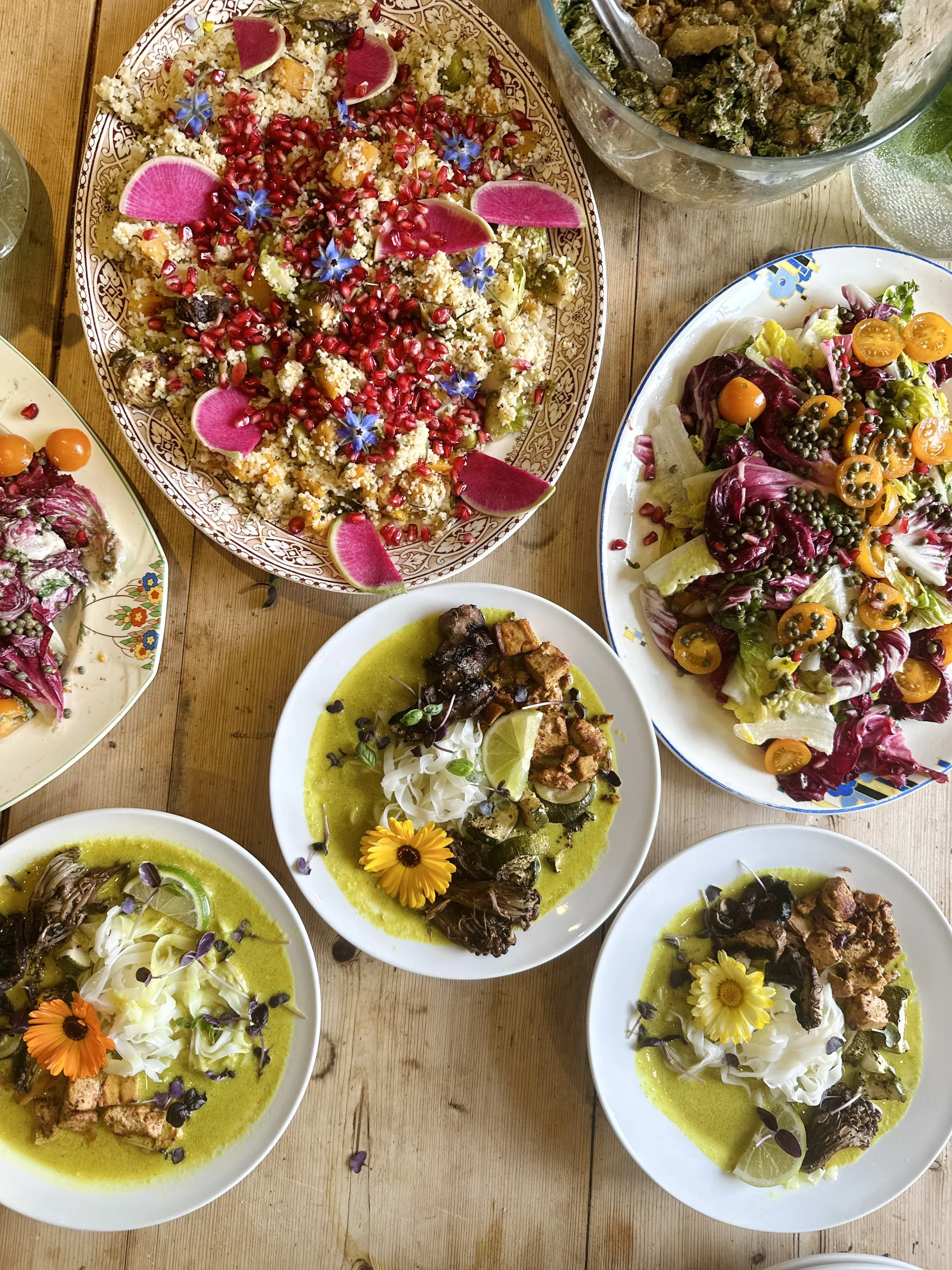 Top-down view of a wooden table with various dishes, including salads with colorful vegetables, a quinoa dish garnished with pomegranate seeds, edible flowers, and watermelon radish slices, and bowls with green curry dishes containing vegetables and 